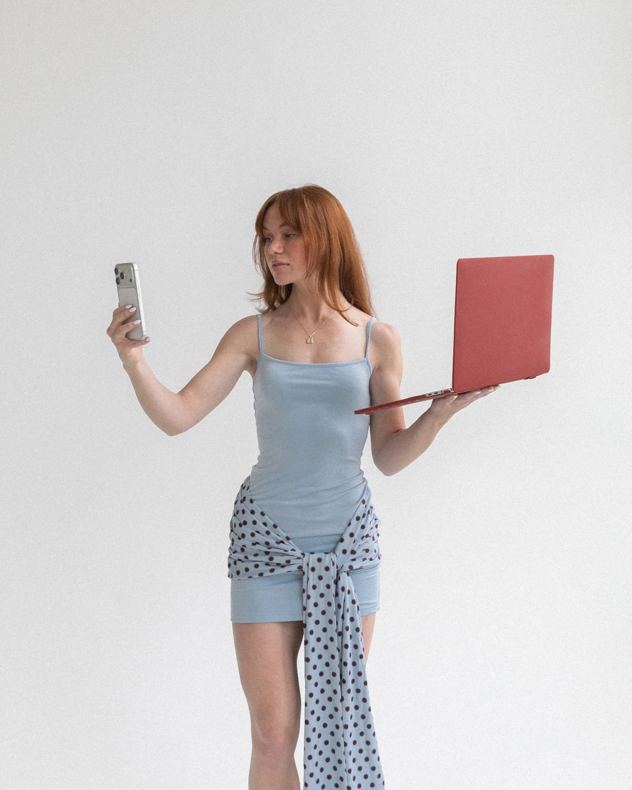 A woman with red hair, wearing a light blue tank top and a polka dot shirt tied around her waist, holds a smartphone in one hand and a pink laptop in the other, standing against a plain white background.