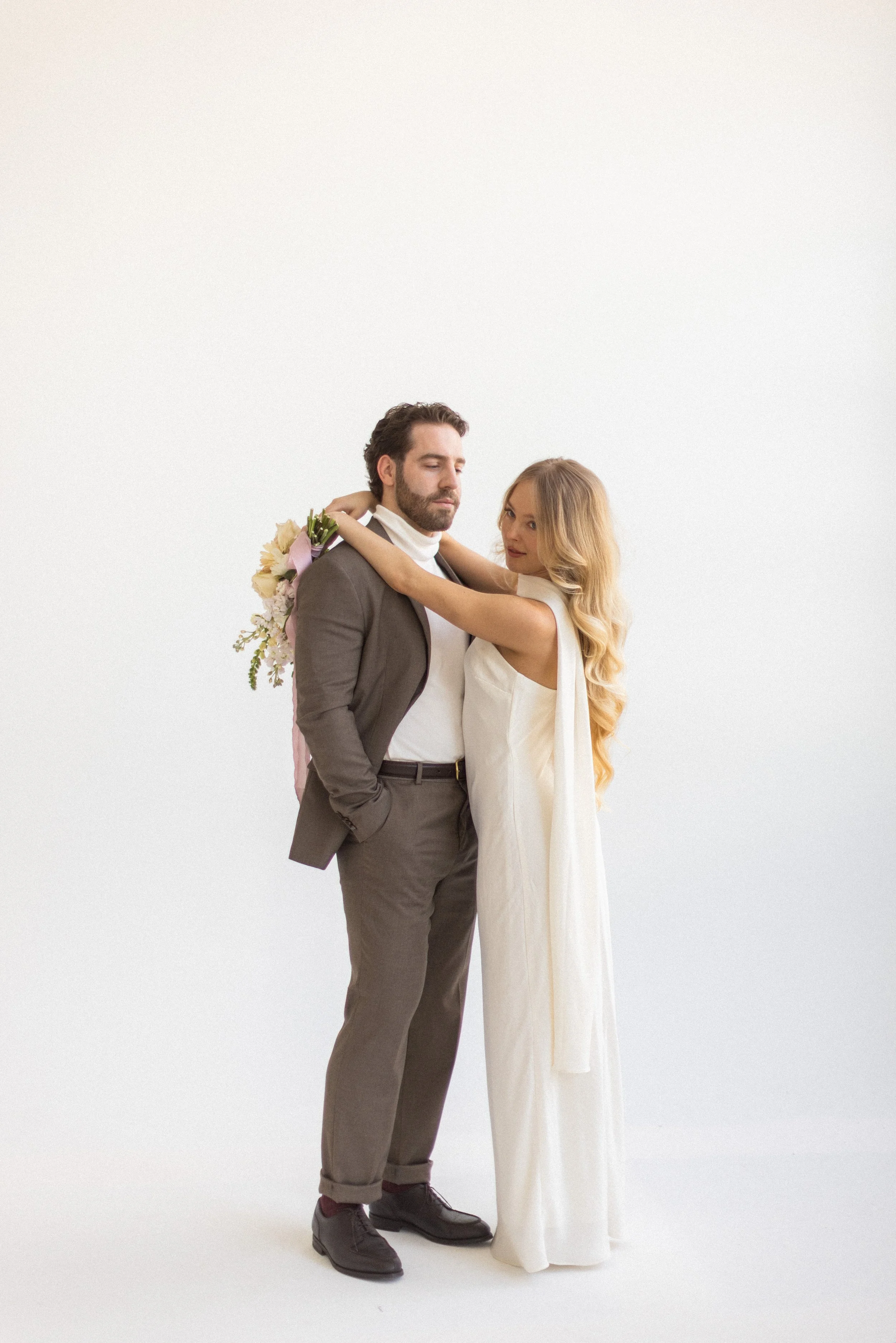 A man and woman stand closely together, with the woman wrapping her arms around the man's neck. The man has a beard, dark hair, and is dressed in a gray suit with an open collar shirt. He carries a bouquet of flowers on his shoulder. The woman has lo