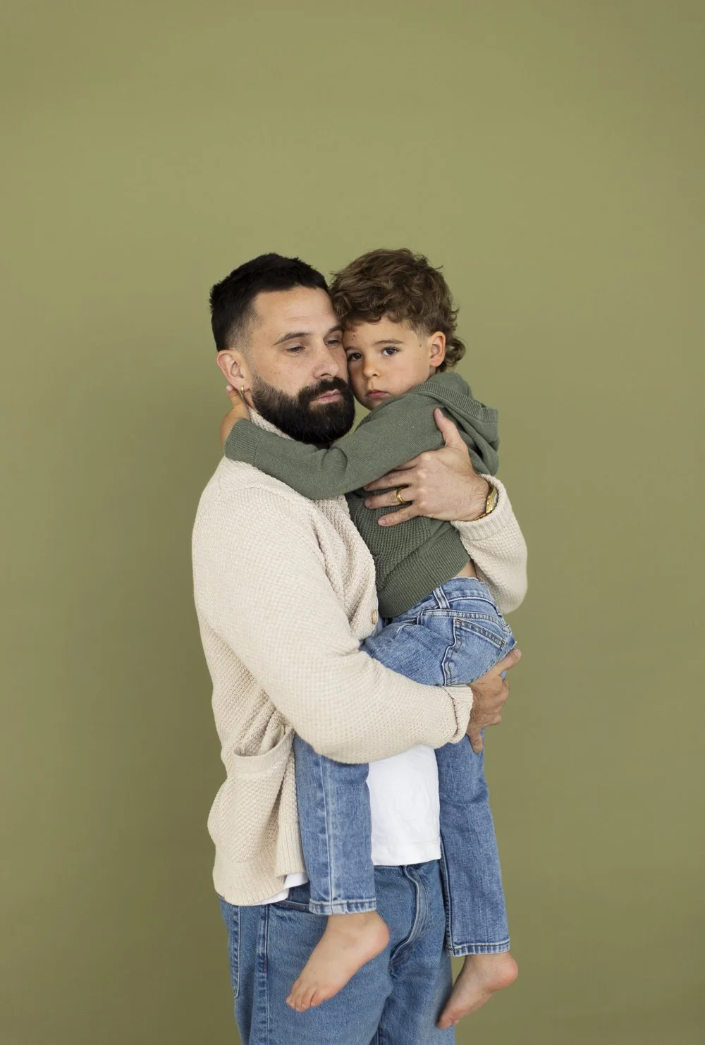 A man with a beard holding a young boy in his arms, both looking at the camera against a plain green background.