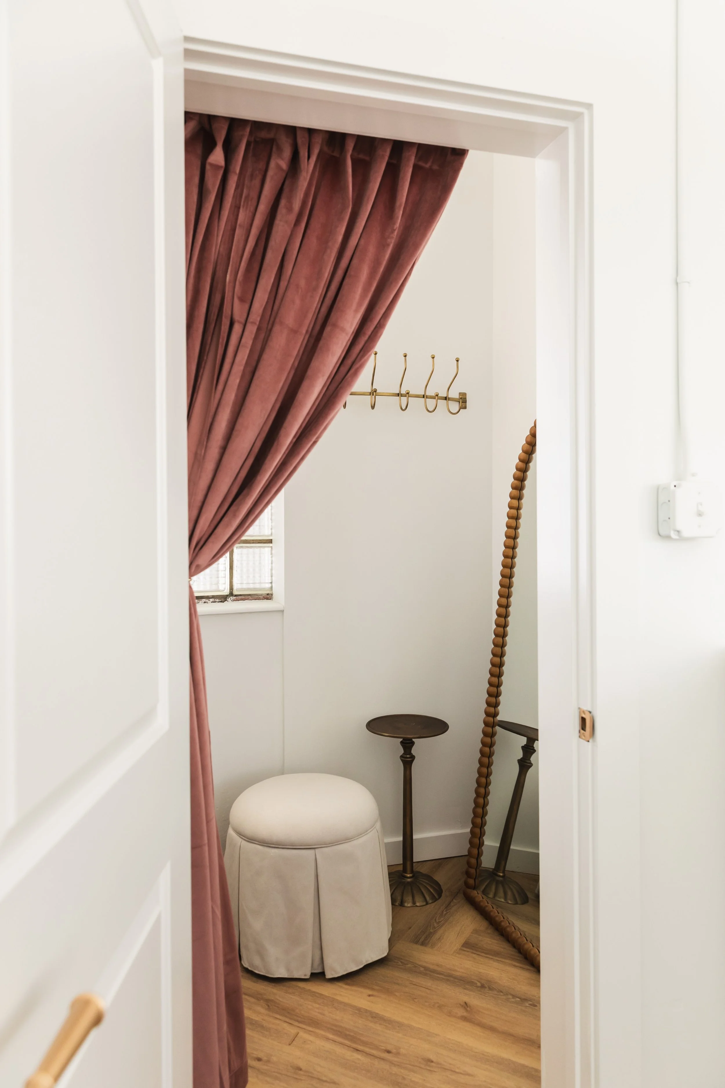 A small room with a wooden floor, a cream-colored stool, a brass wall hook rack, a mirror leaning against the wall, a wooden bead curtain, and a window with pinkish-brown curtains.