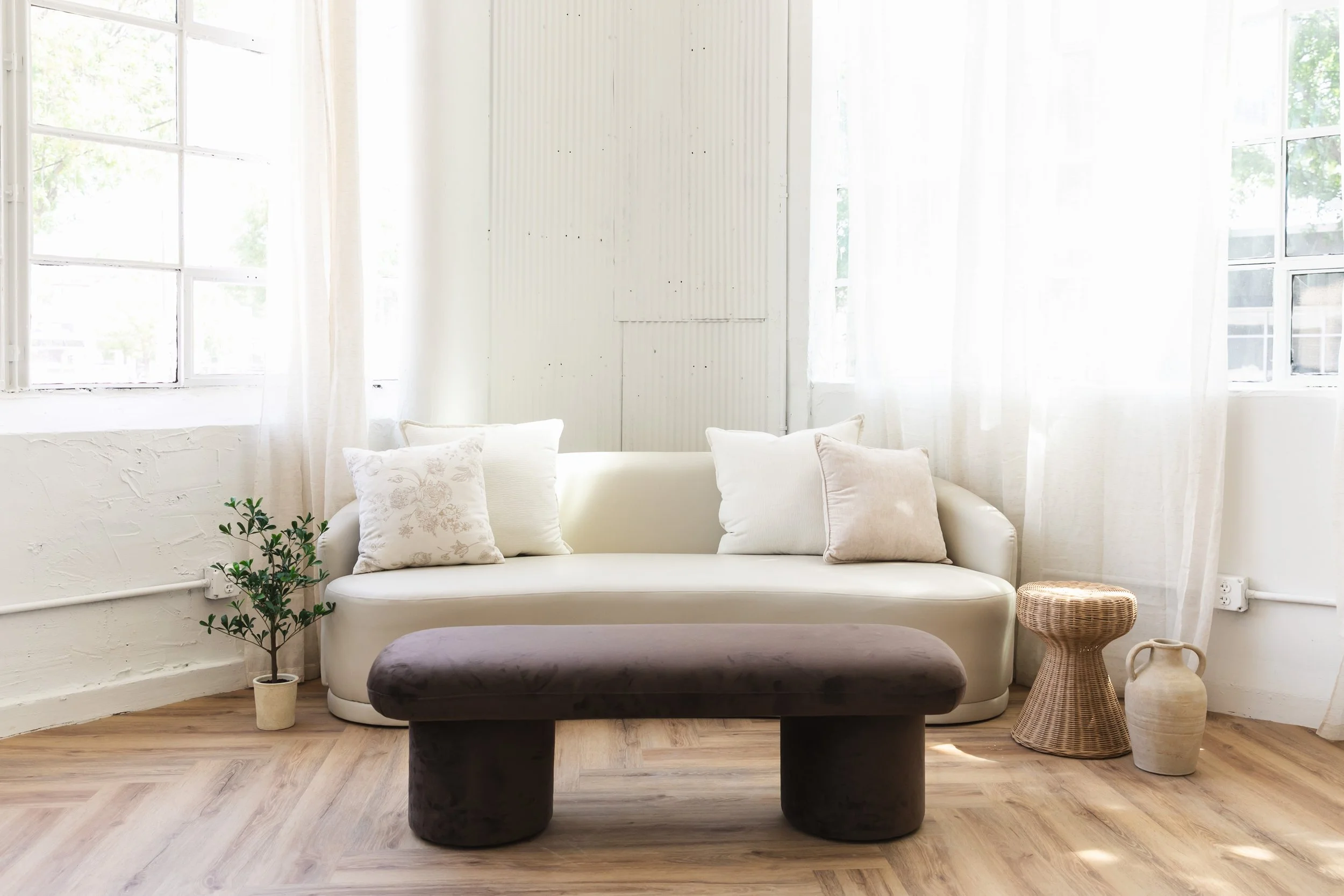 Bright living room with white sofa, beige and white pillows, a dark upholstered ottoman, a potted plant, and woven side tables by large windows with white curtains.