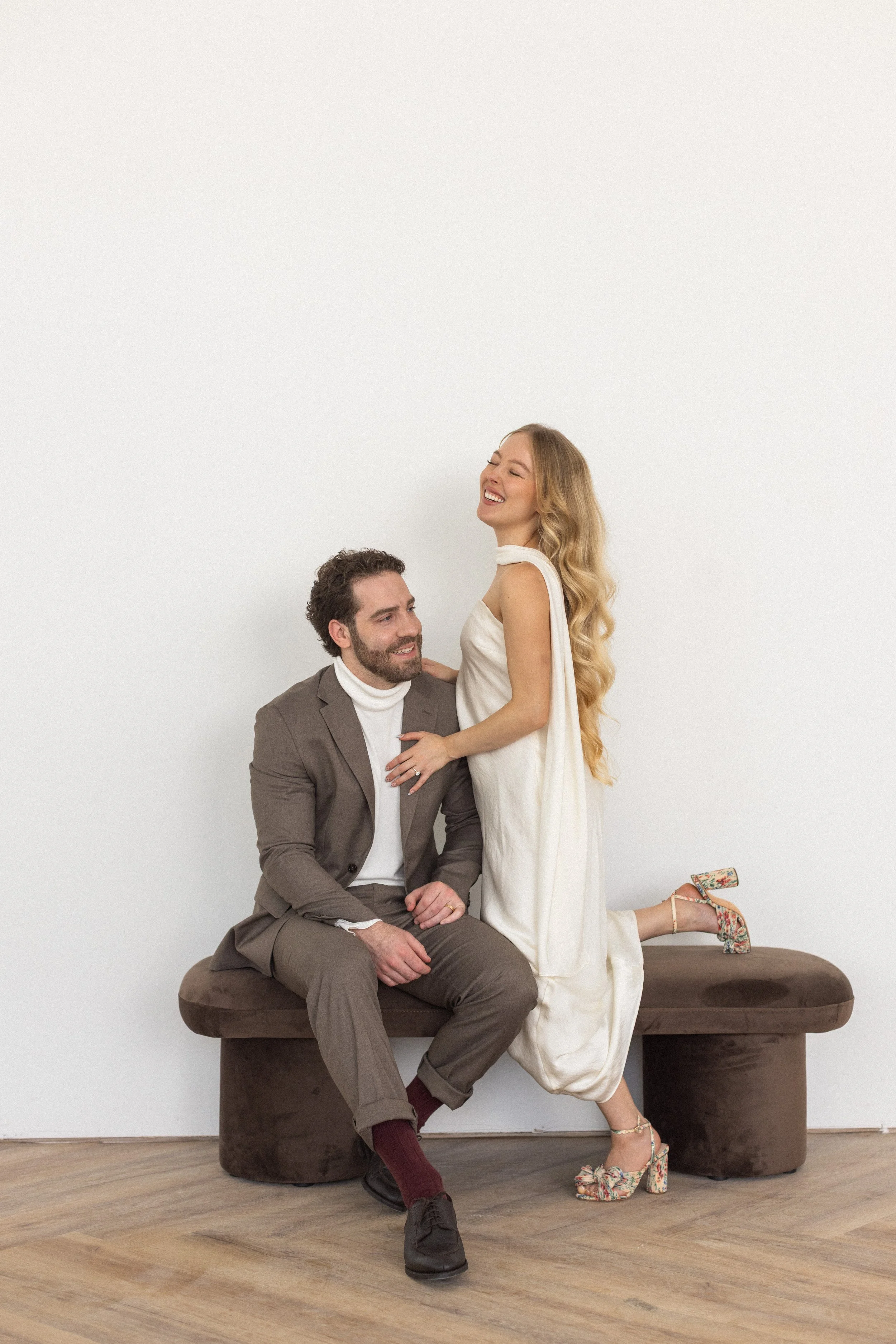 A man and a woman are sitting and standing on a brown bench against a plain white wall. The man is dressed in a brown suit with a white turtleneck, and the woman is wearing a cream-colored dress with floral heels. The woman is smiling and leaning on 