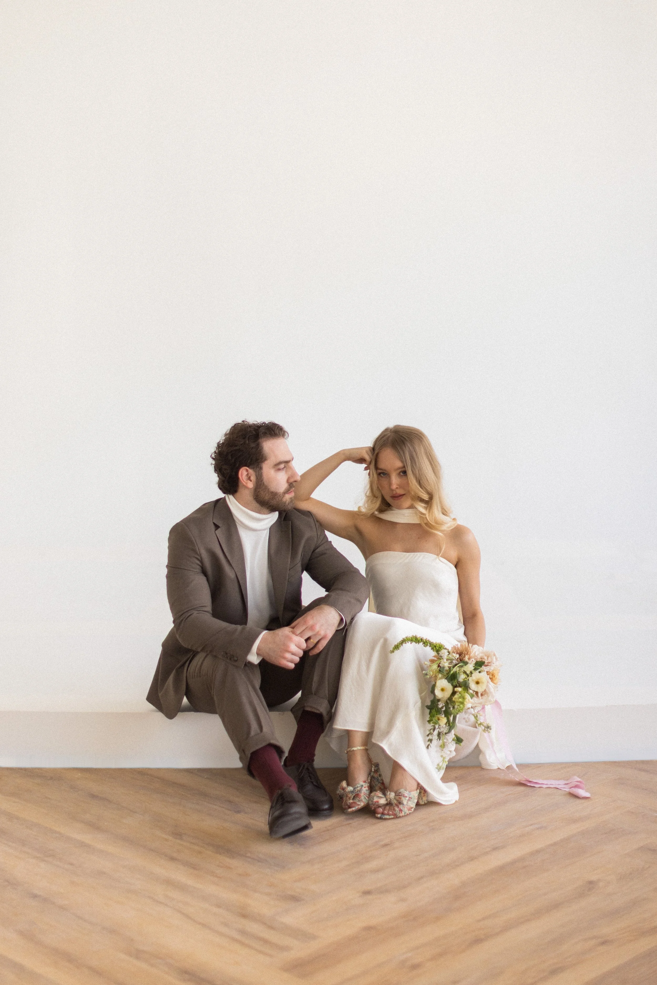 A man and woman sitting on the floor against a white wall. The woman is in a white dress holding a bouquet and wearing floral heels, while the man is in a brown suit with a white turtleneck and dark shoes.