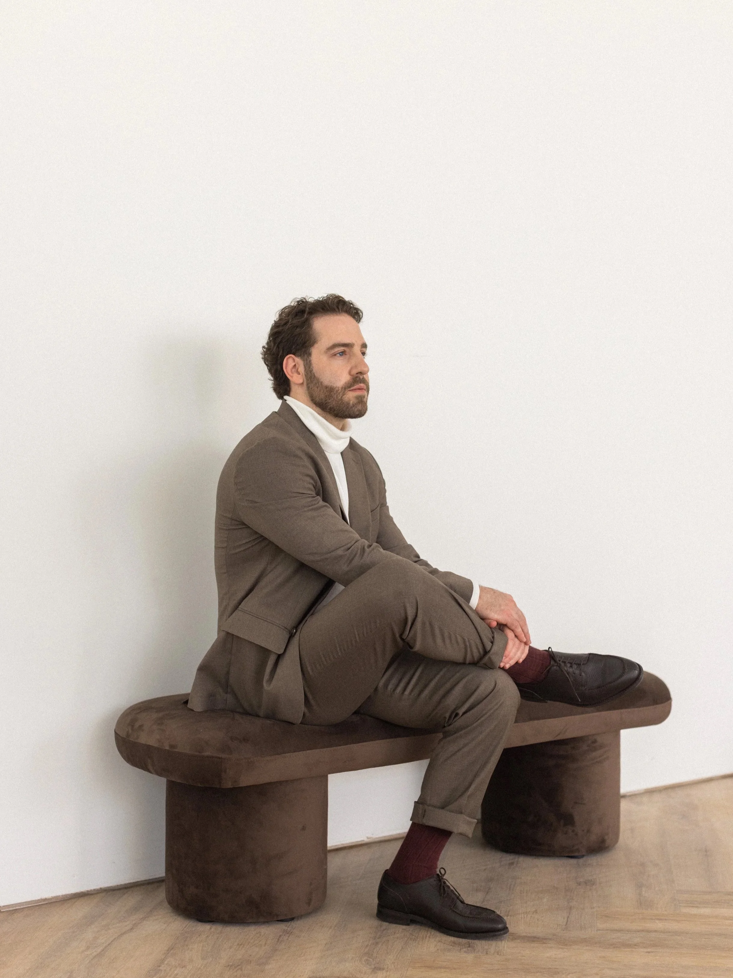 A man in a brown suit and white turtleneck sitting on a brown bench with his legs crossed, against a white wall.