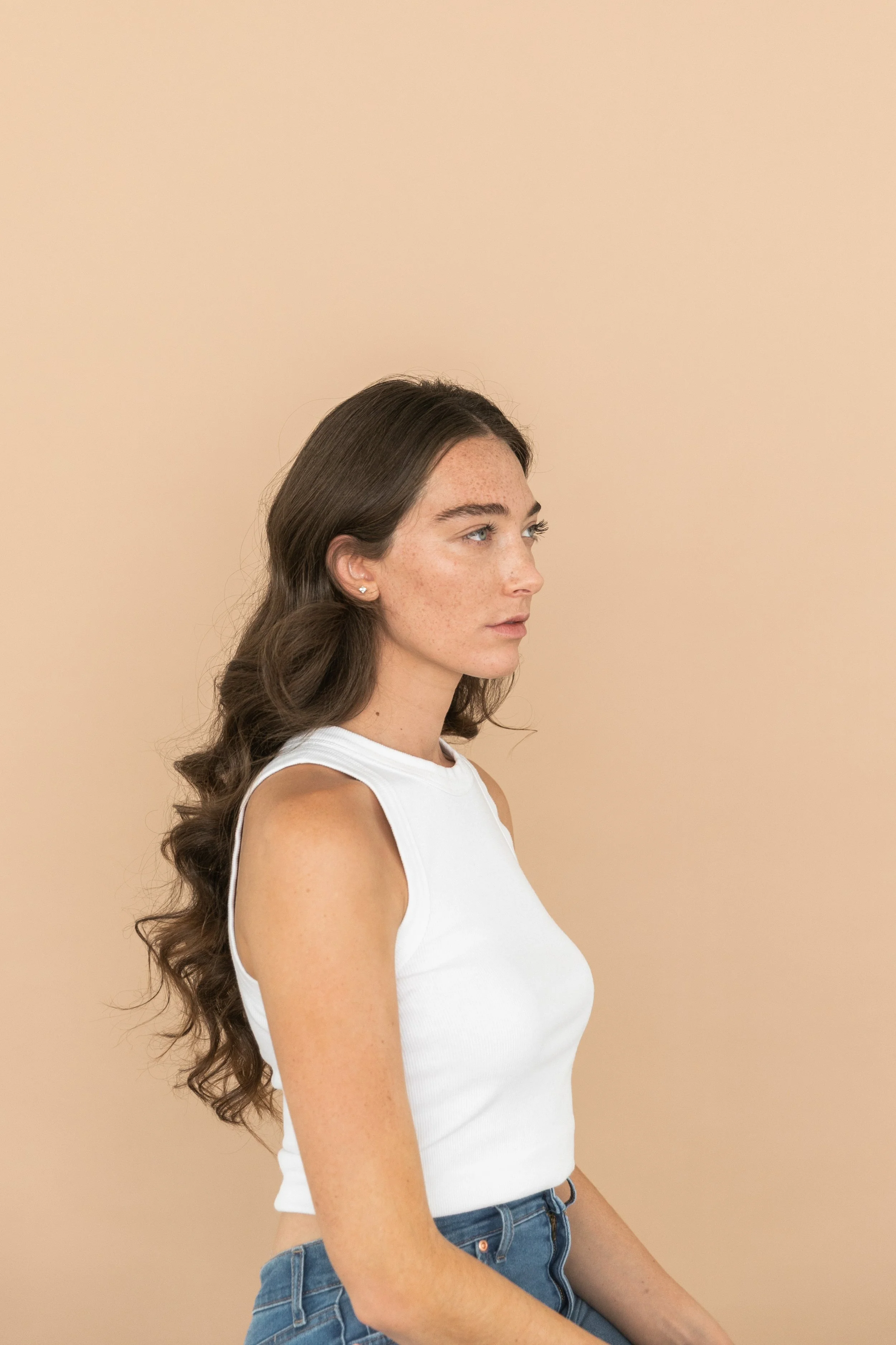 Portrait of a young woman with long wavy brown hair, wearing a white sleeveless top and blue jeans, against a beige background.