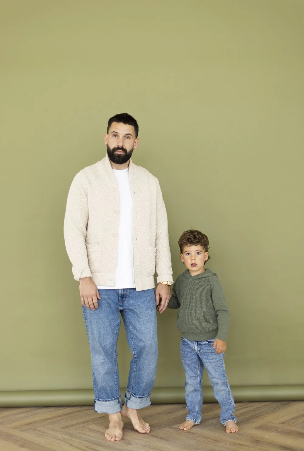 A man and a young boy standing barefoot in front of a plain green wall. The man has dark hair, a beard, and is wearing a cream-colored cardigan and blue jeans. The boy has curly hair, and is wearing a green hoodie and blue jeans.