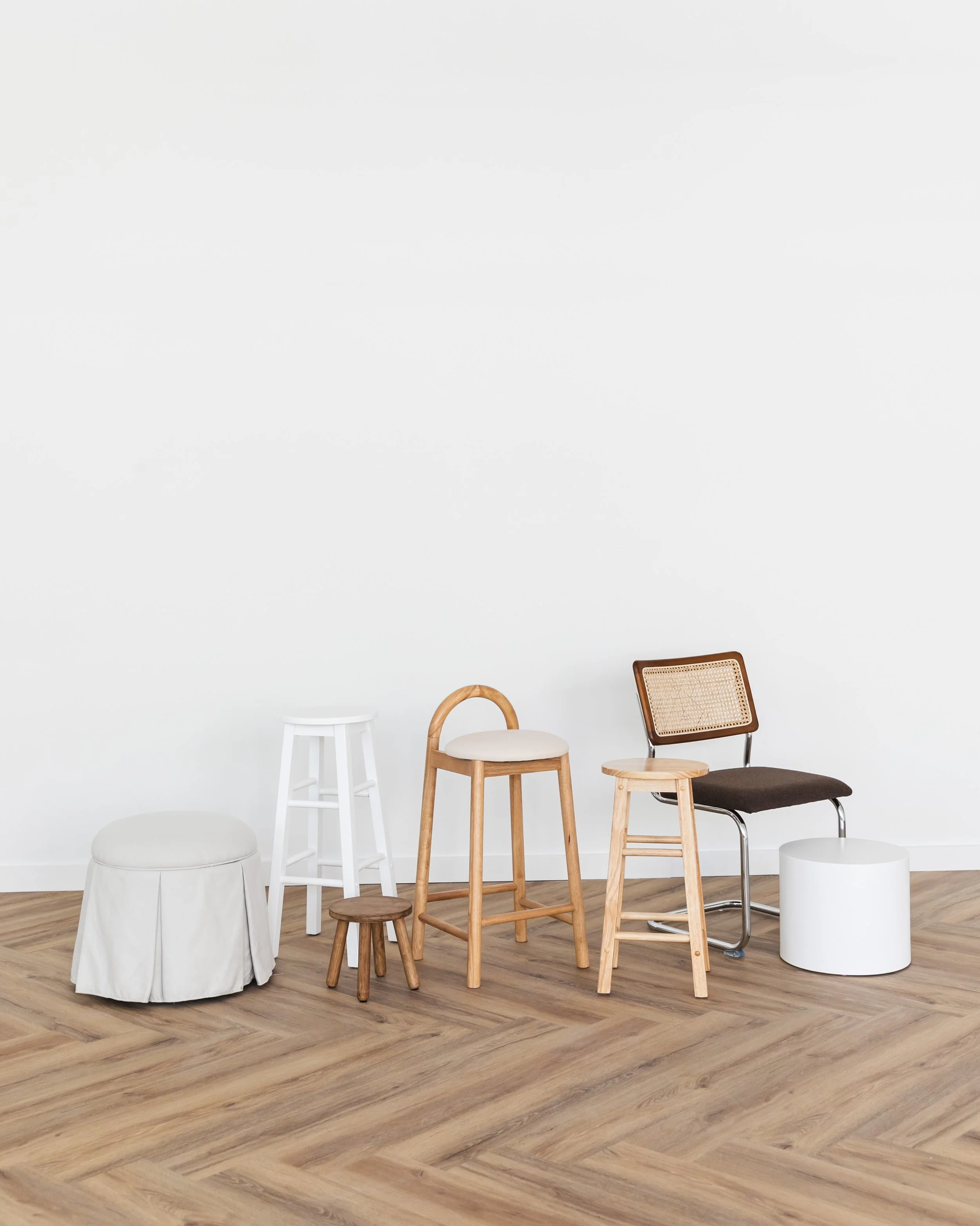 Various chairs and stools lined up against a white wall on a wooden floor.