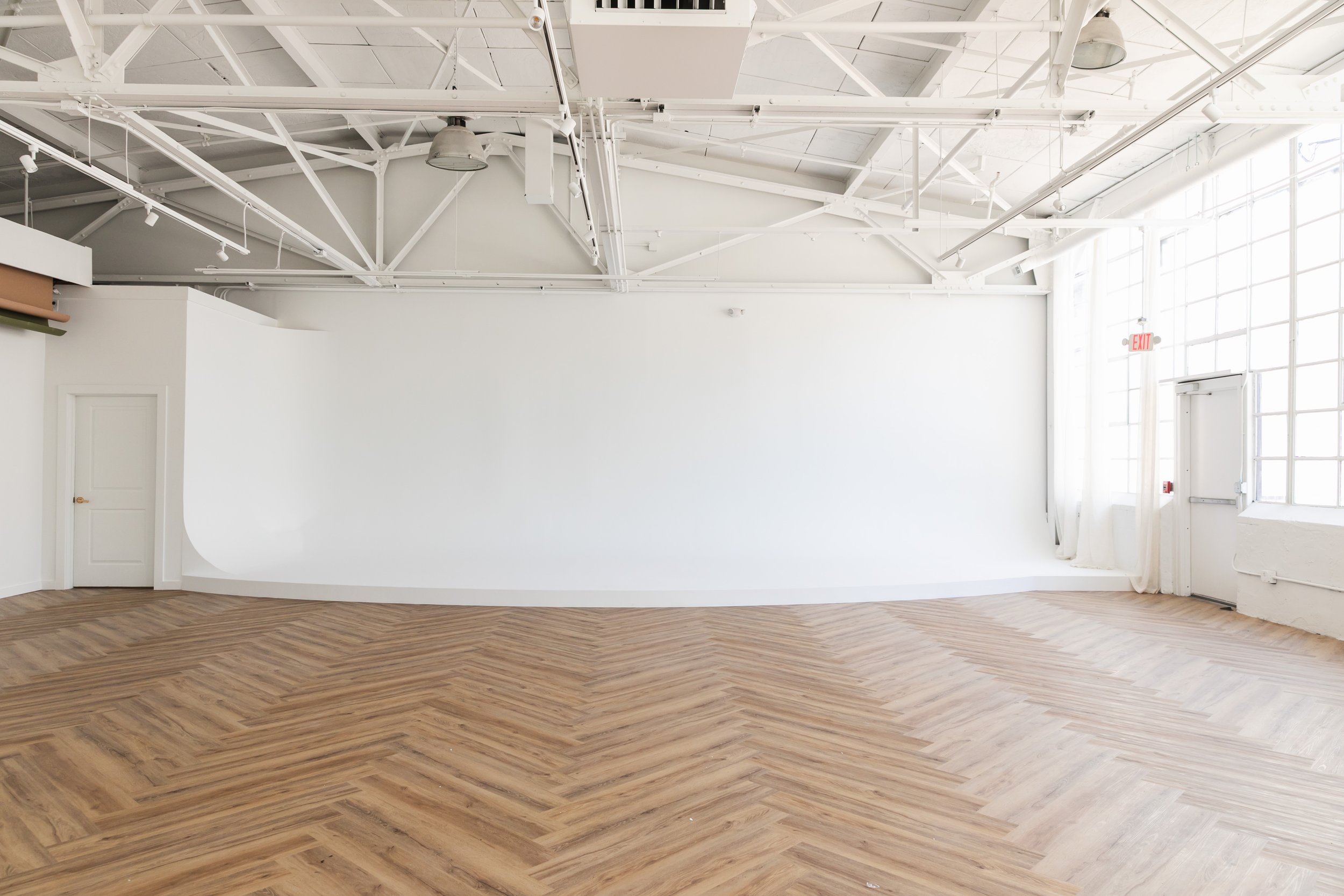 Empty white room with large windows, wooden chevron patterned floor, white curtain, and visible ceiling beams.