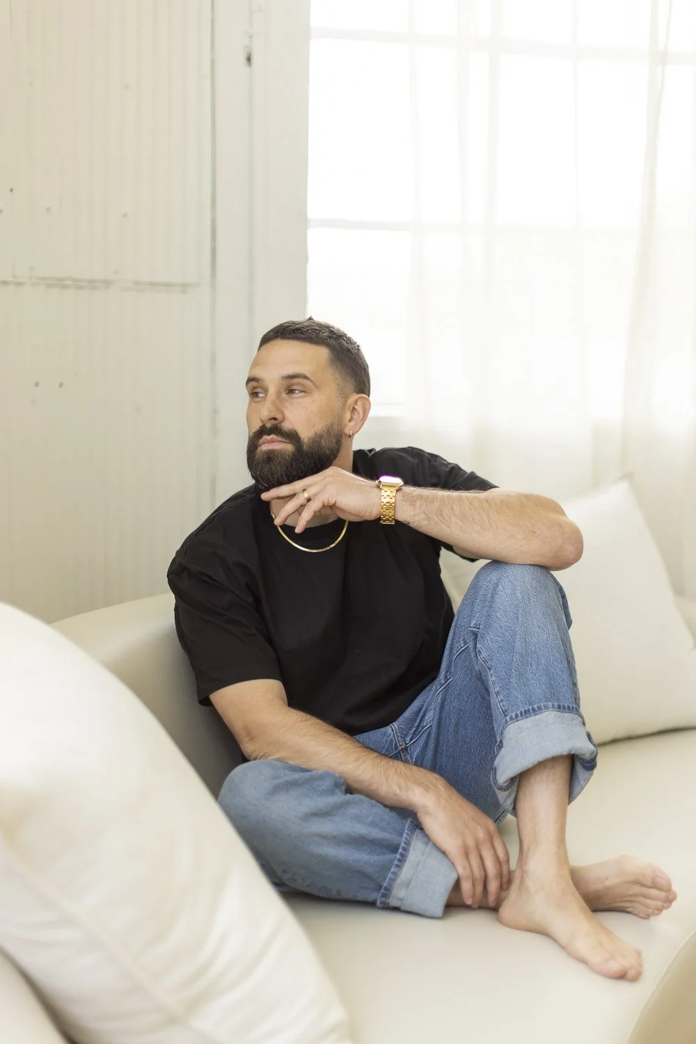 A bearded man with short dark hair, wearing a black t-shirt, blue jeans, and a gold watch, sitting on a white sofa in a bright room with large windows, looking thoughtfully to his left.