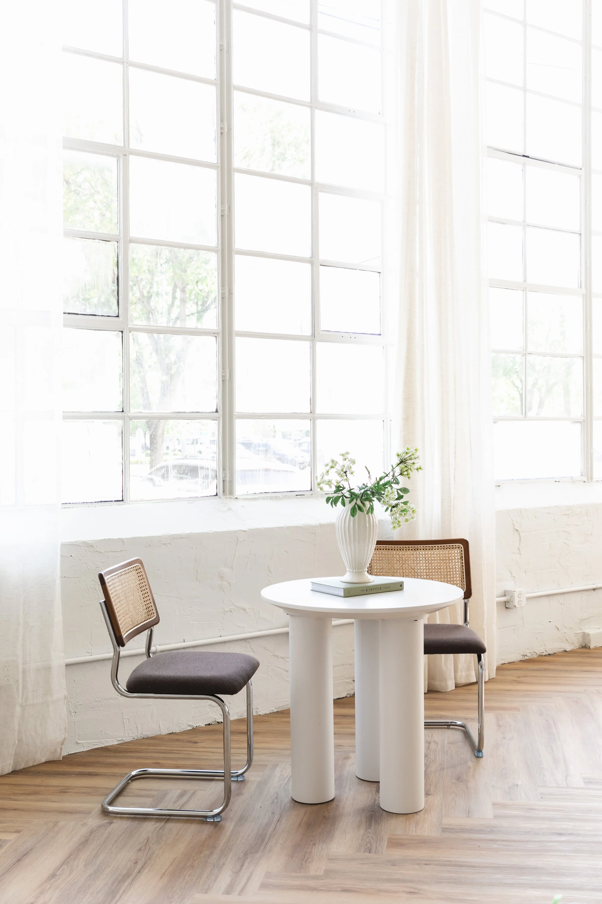 A bright room with large windows, white curtains, a white round table, two chairs, and a vase with green leaves and white flowers on the table.