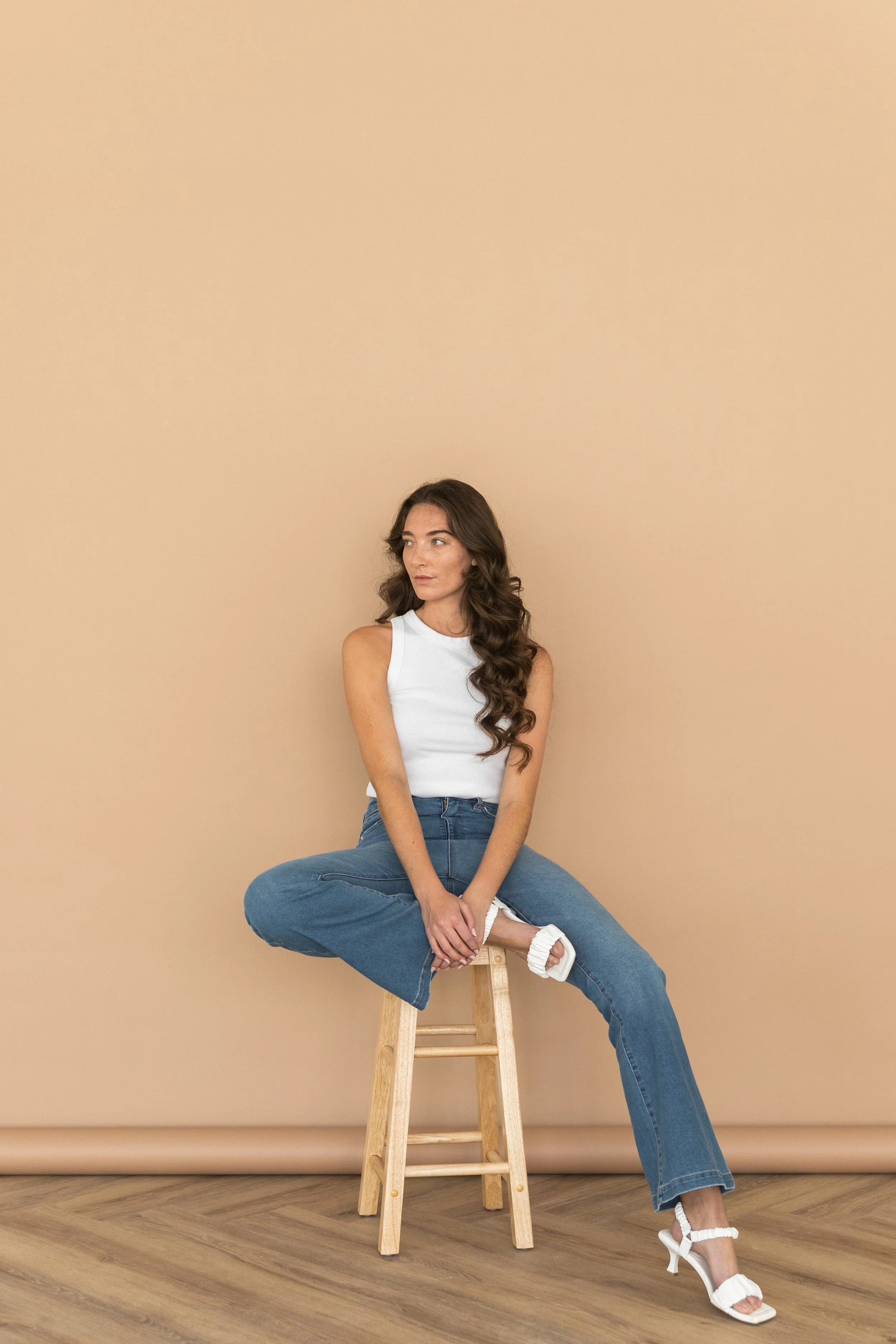 A woman with long, curly brown hair sitting on a wooden stool against a beige background, wearing a white sleeveless top, blue jeans, and white high-heeled sandals.
