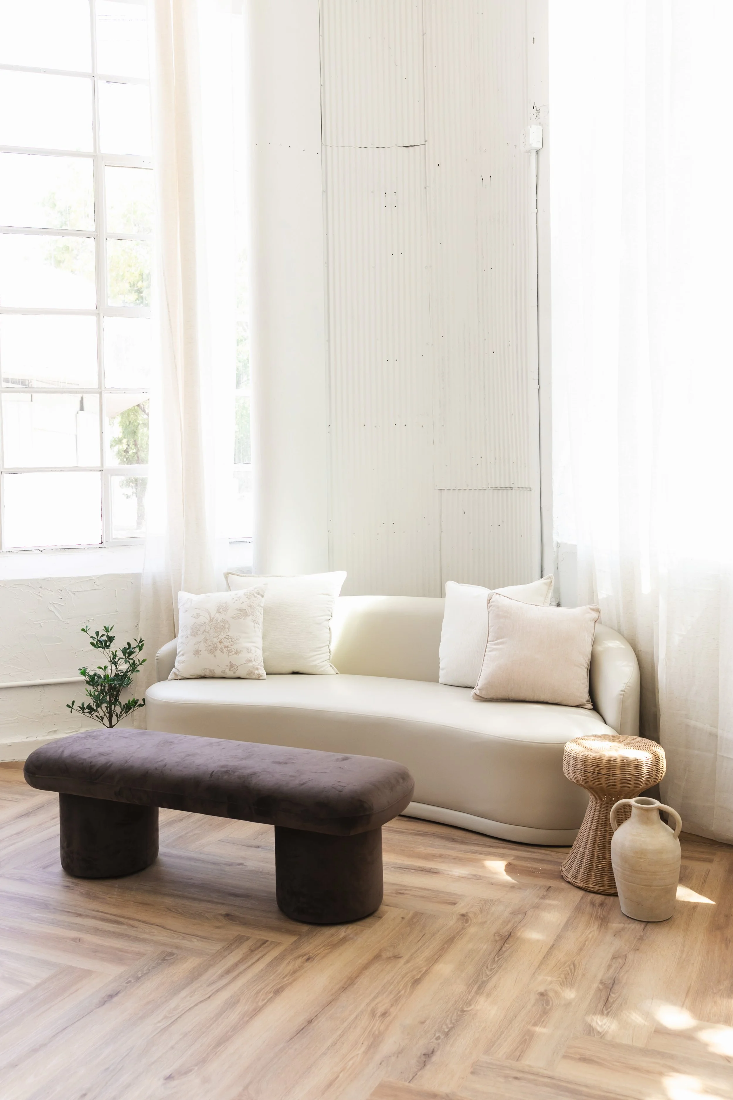 Bright living room with a white sofa, dark velvet bench, wicker side table, ceramic vases, and sheer curtains by large window.