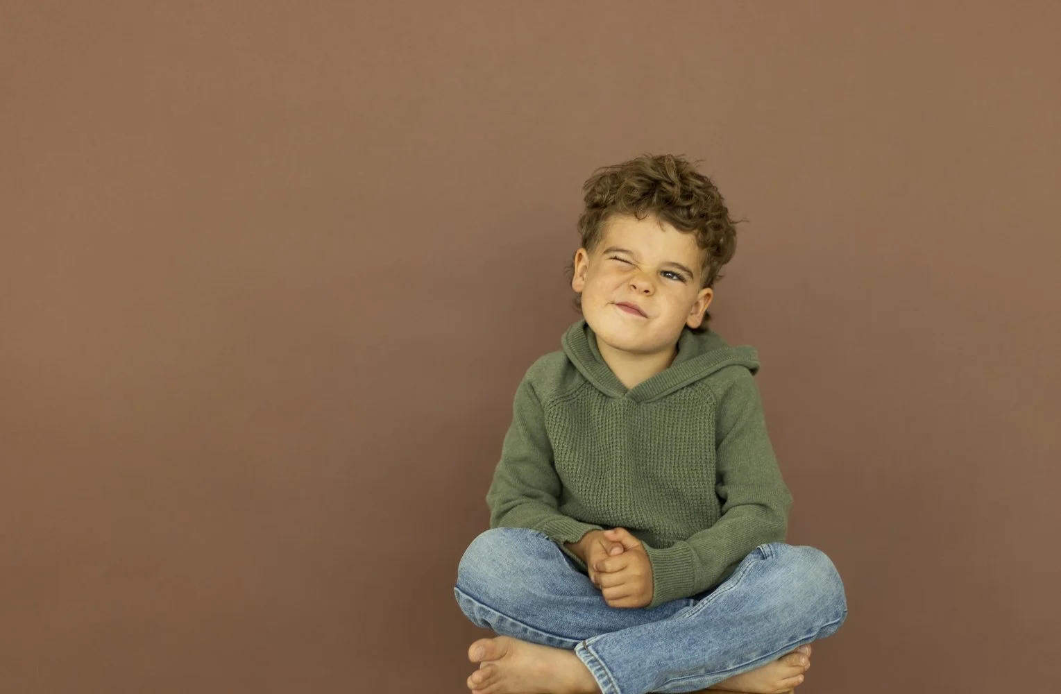 A young boy with curly hair sitting cross-legged against a plain brown background, wearing a green hoodie and blue jeans, making a playful face by squinting one eye.