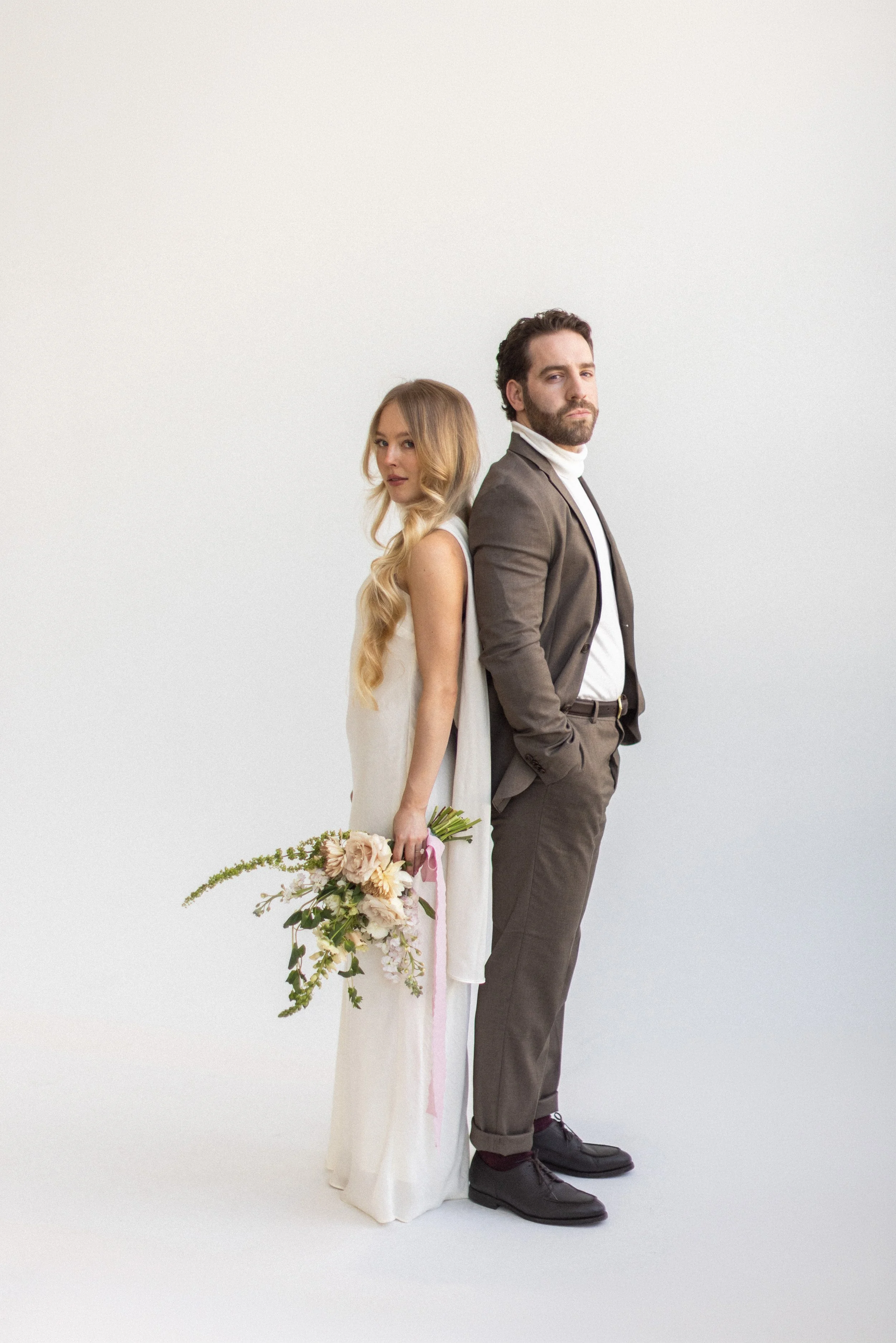 A woman in a white dress holding a bouquet of flowers and a man in a gray suit standing back to back against a plain white background.