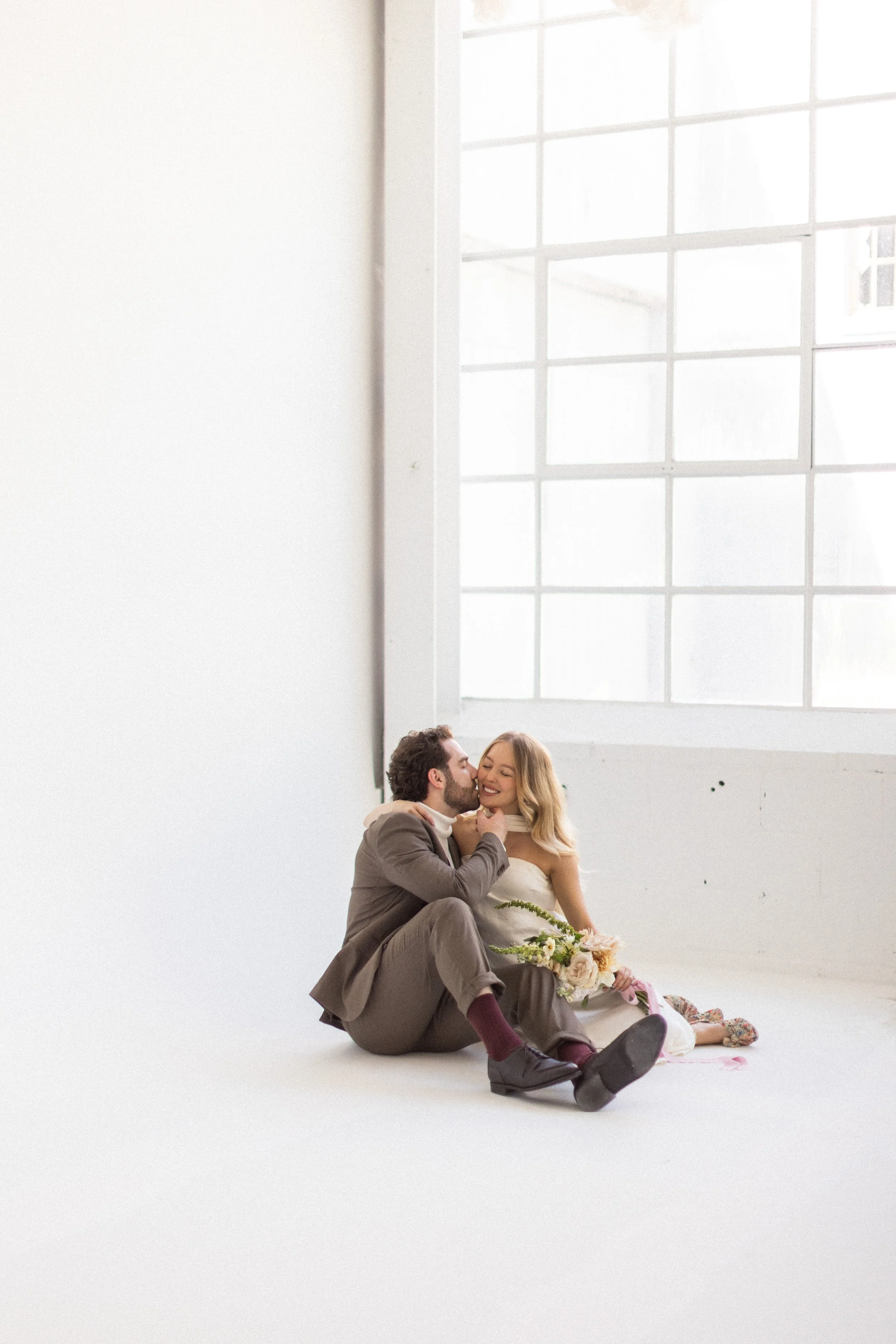 A couple sitting on the floor, smiling and embracing in a bright, minimalistic room with large windows, the woman holding a bouquet of flowers.