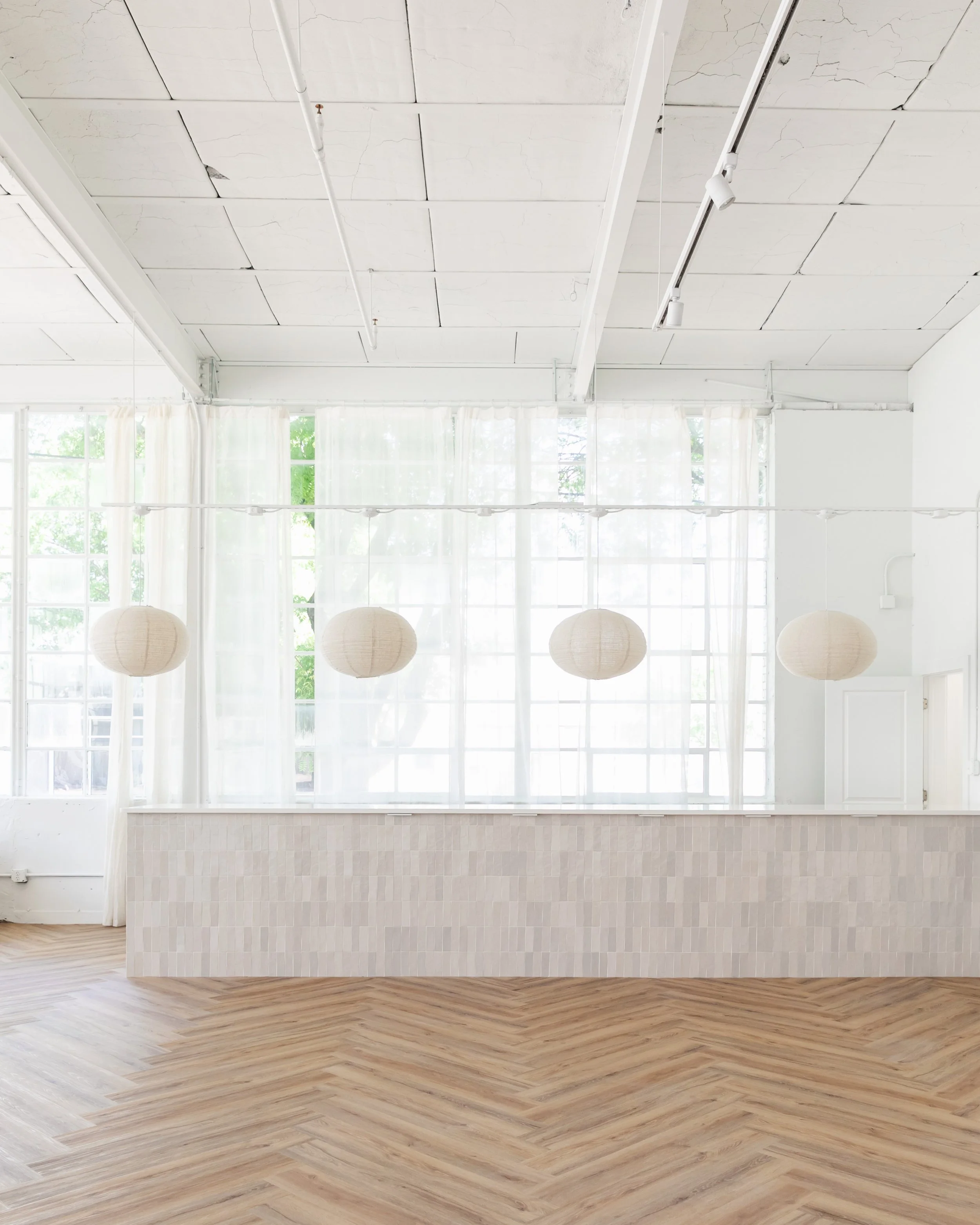 Bright room with large windows and sheer curtains, hanging paper lanterns, a tiled half wall, and a herringbone-patterned wooden floor.