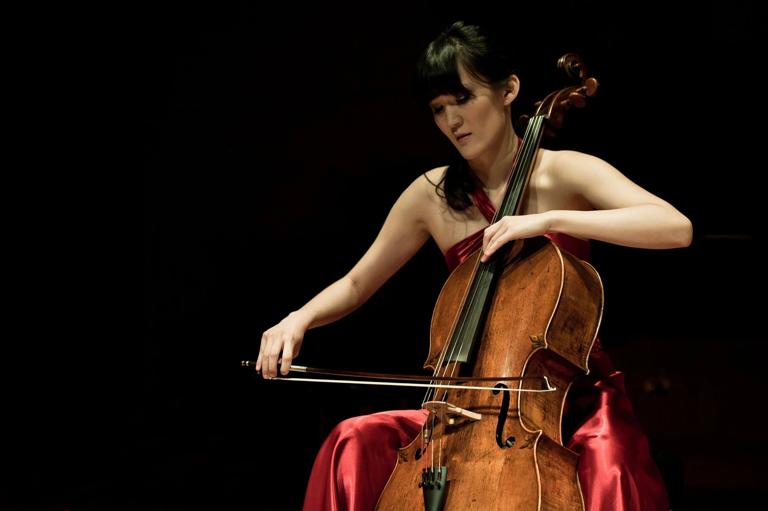 Mimi Yu is in a red dress playing a cello on stage against a black background.