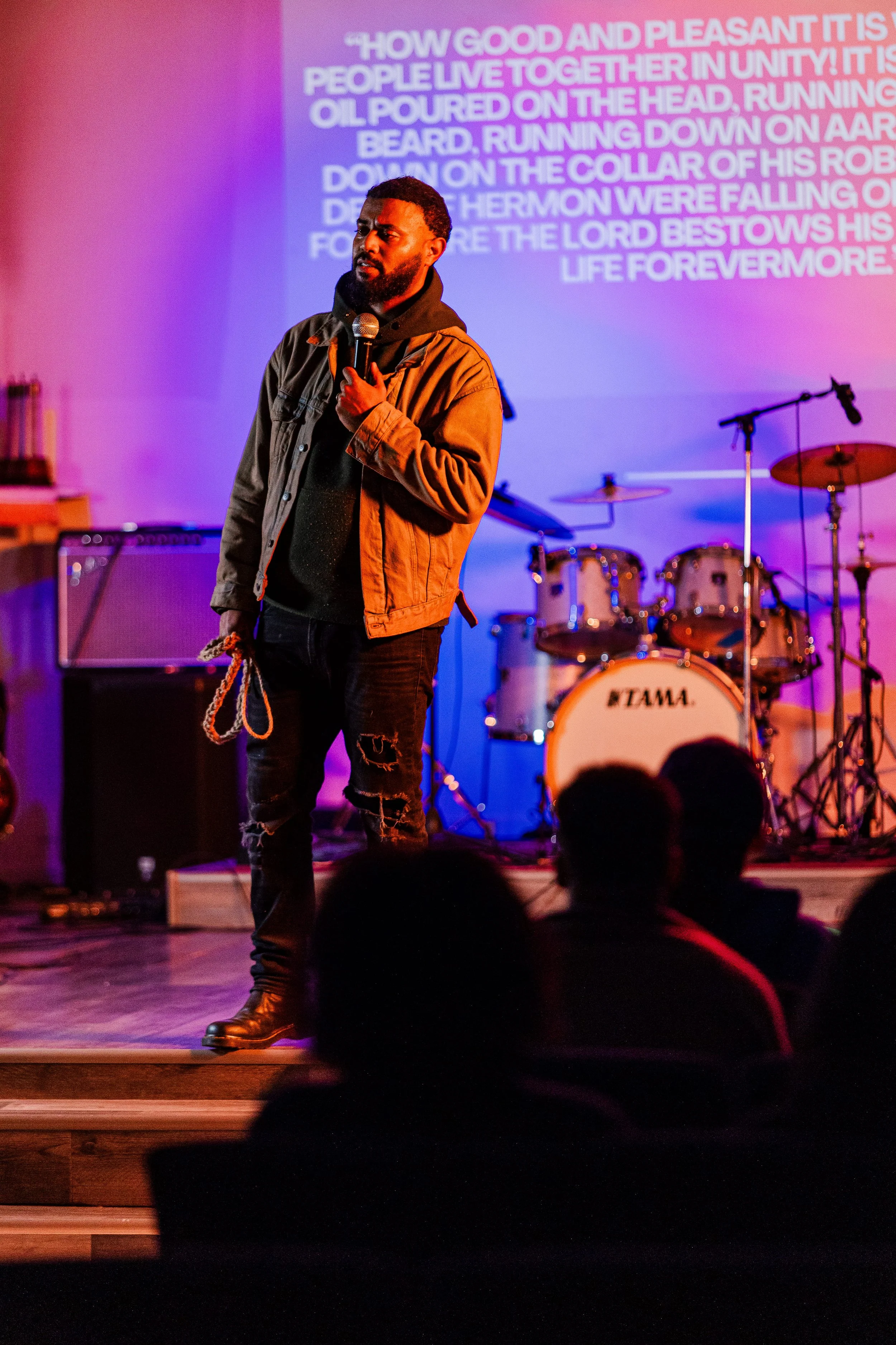 A man standing on a stage holding a microphone, with musical drums and equipment in the background, and a projected text on the wall.