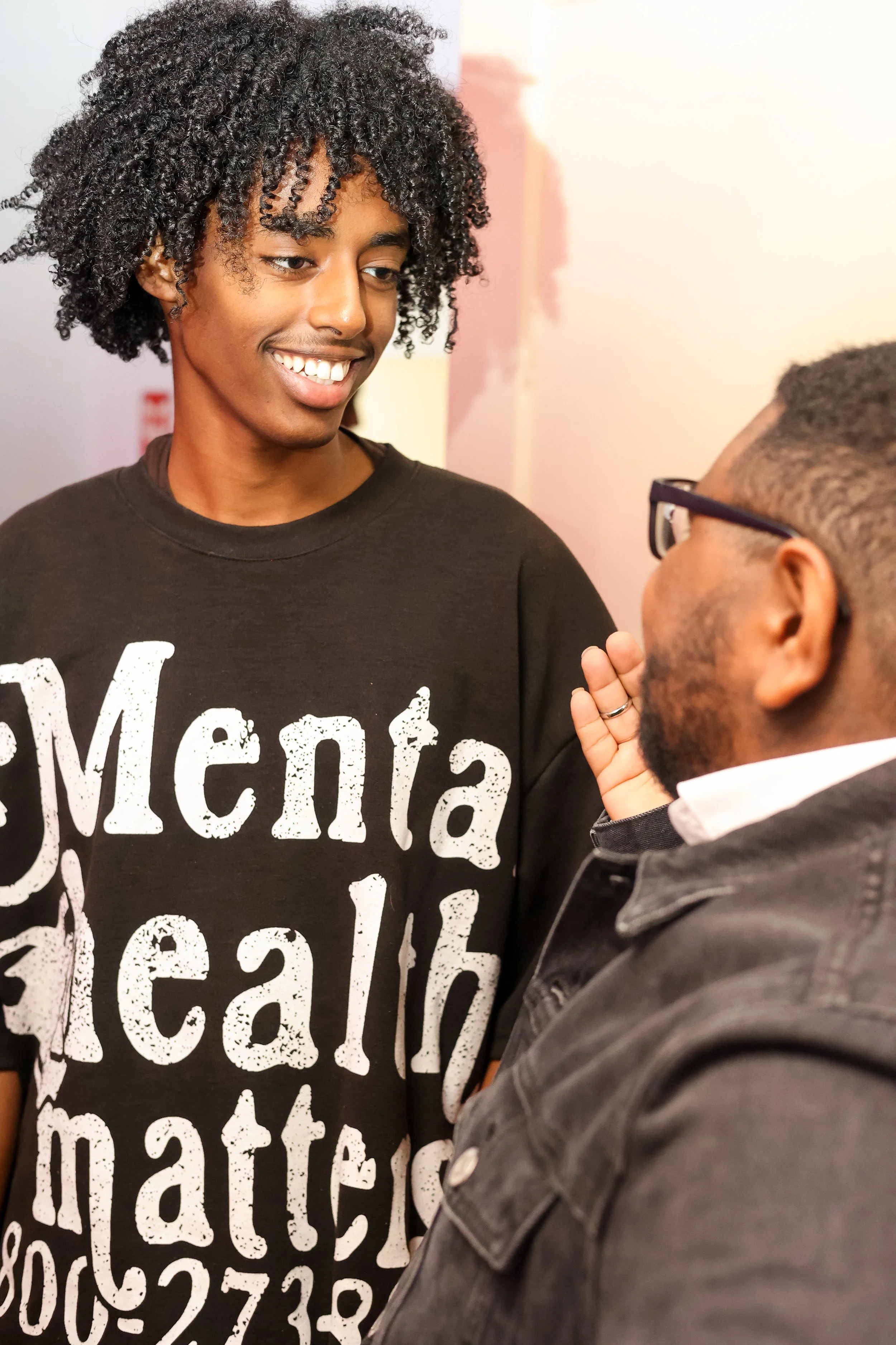 A young man with curly hair smiling and talking to a man with glasses, both engaged in conversation, indoors.