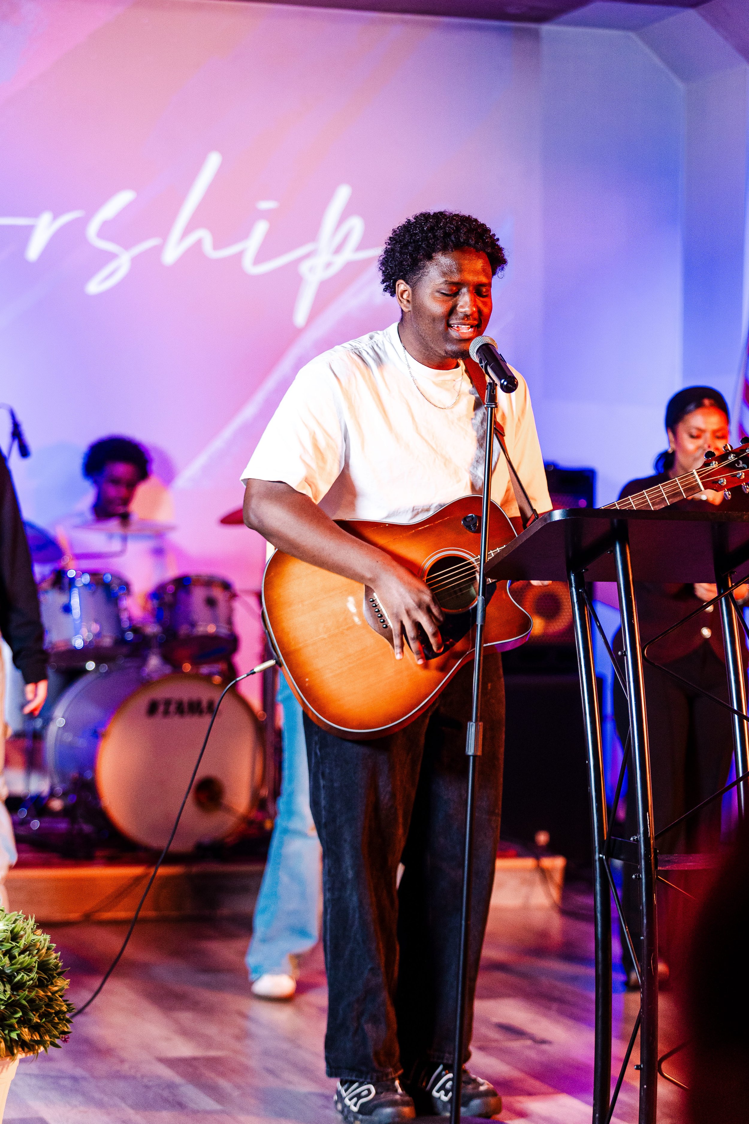 A man singing and playing an acoustic guitar during a live music performance, with a band playing drums and other musicians in the background, on a stage with colorful lighting.