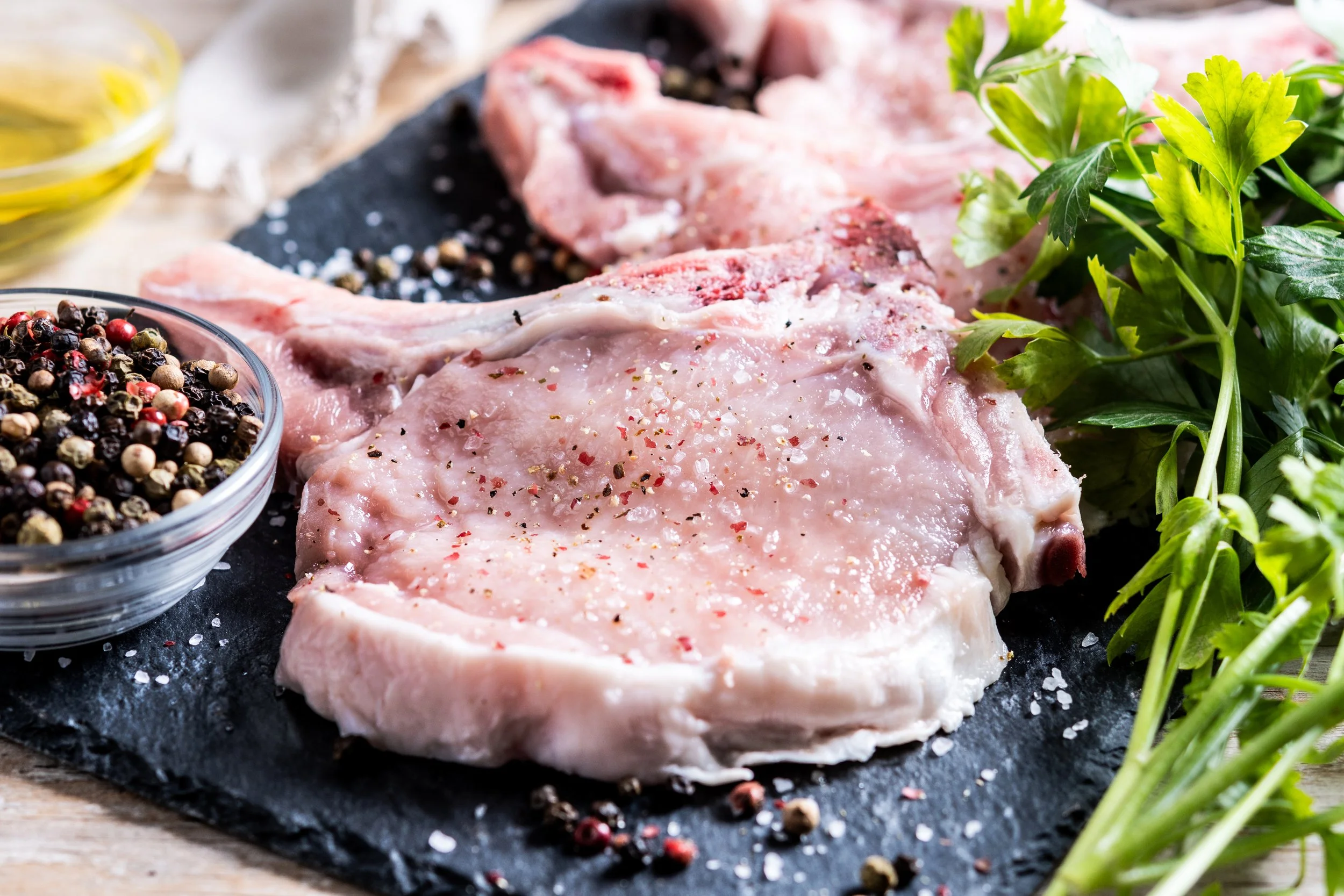 Raw pork chops seasoned with salt and pepper on a black slate board, surrounded by fresh parsley, a bowl of mixed peppercorns, and a small bowl of olive oil.