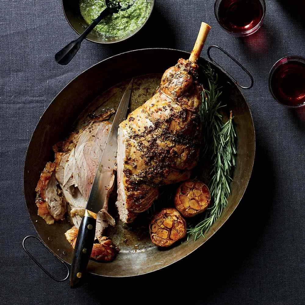 Roast leg of lamb garnished with rosemary, served with roasted garlic cloves, with a side of green sauce and two glasses of red wine on a dark table.