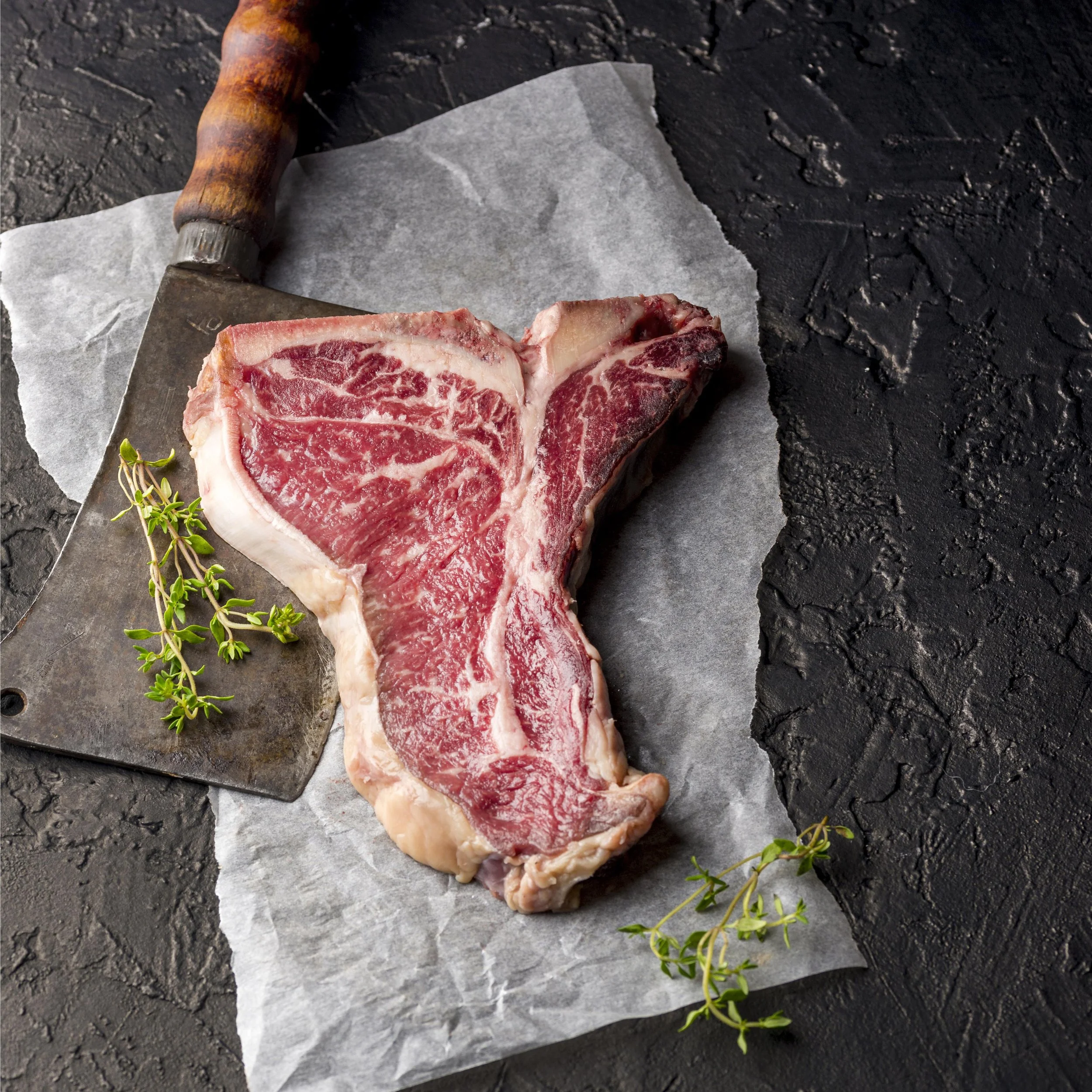 Raw T-bone steak on parchment paper with sprigs of thyme on a dark textured surface.