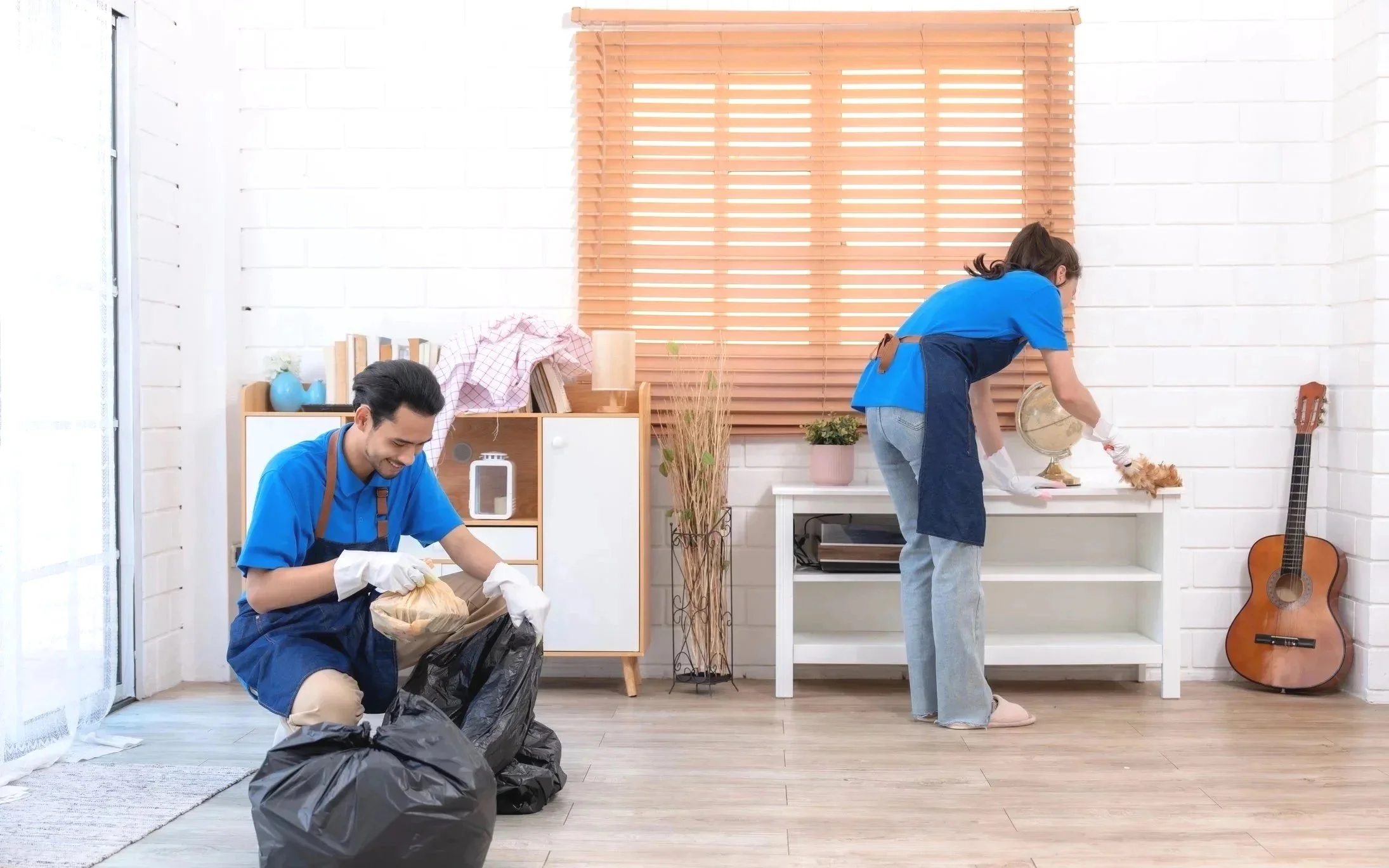 Two people wearing blue shirts and aprons are cleaning and organizing a room. One person is crouched down, placing items into a trash bag, while the other is standing, dusting or wiping an object on a white table. The room has a white brick wall, a window with wooden blinds, and a guitar leaning against the wall.