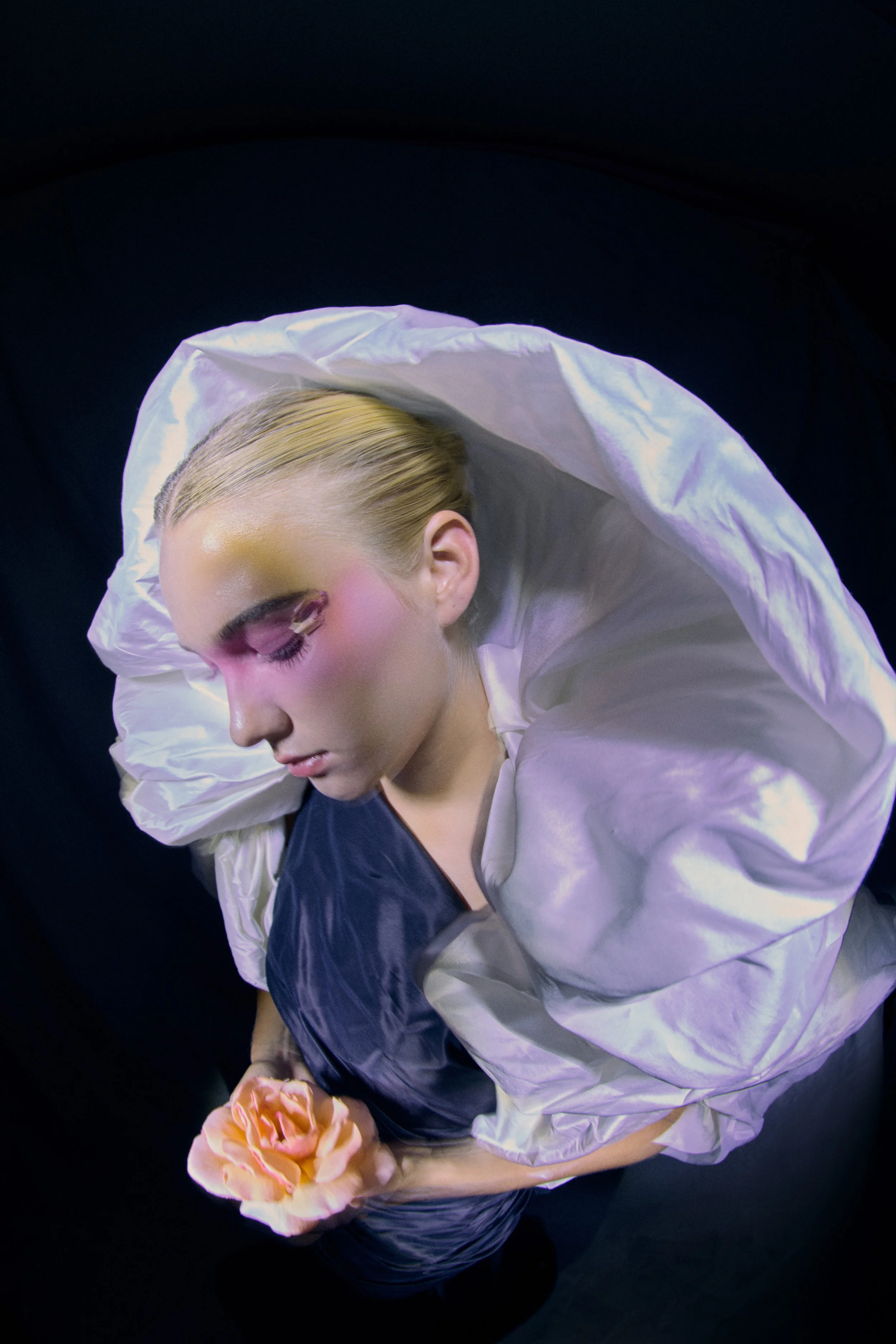 A woman with blonde hair styled back, wearing makeup with pink eyeshadow, dressed in a black top and silver jacket, holding a peach-colored flower, against a black background.