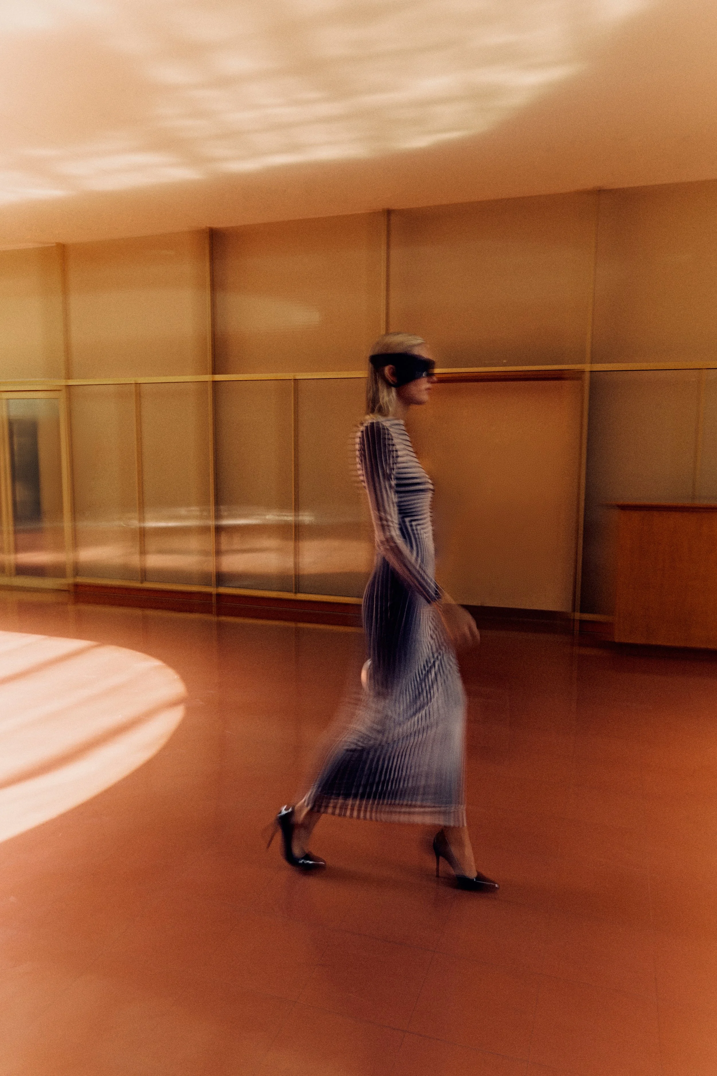 A woman walking in a stylish striped dress and high heels, wearing a blindfold, in a room with a wooden wall and warm lighting.