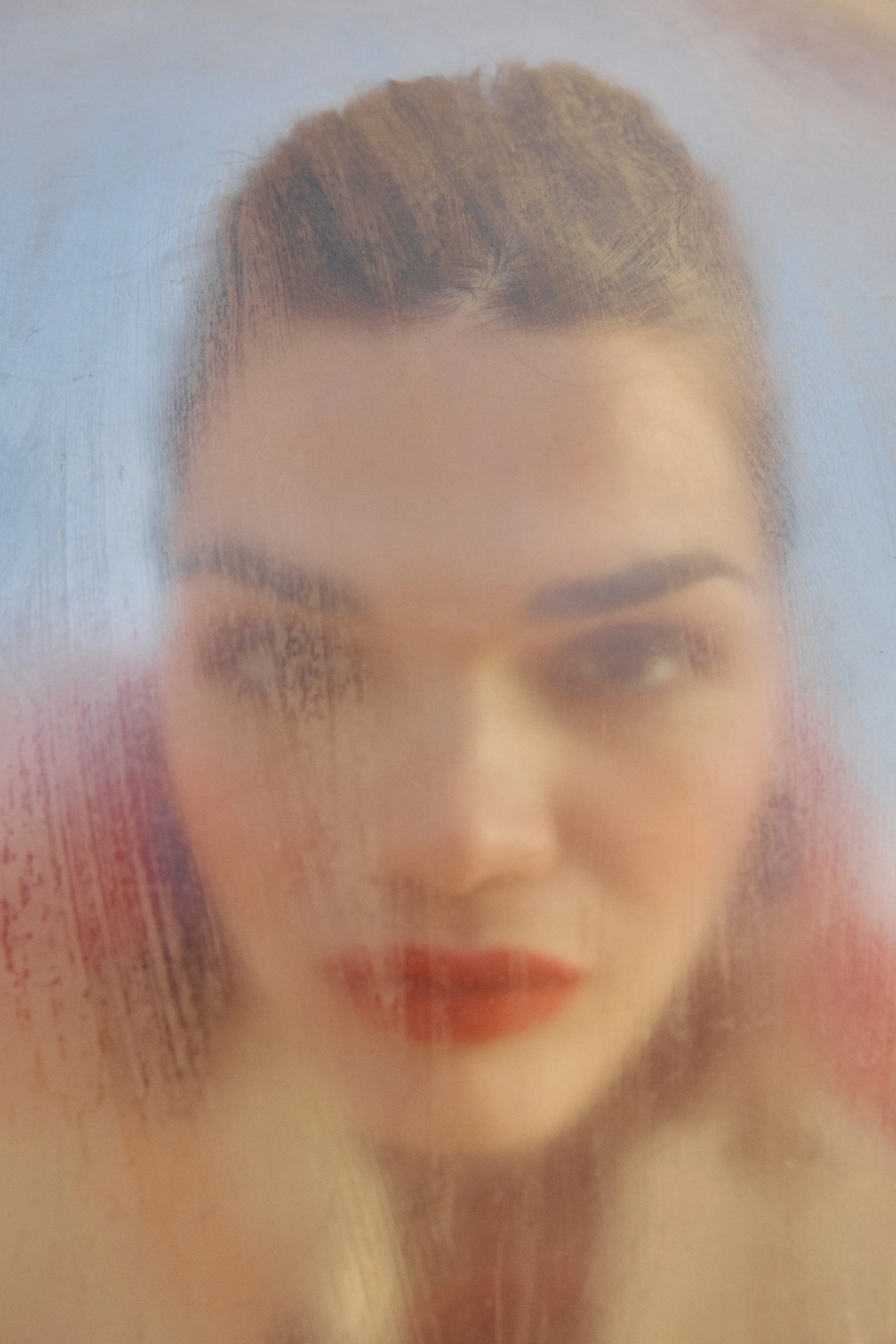 Close-up of a woman's face visible through a frosted glass surface, with only her lips and part of her forehead visible.