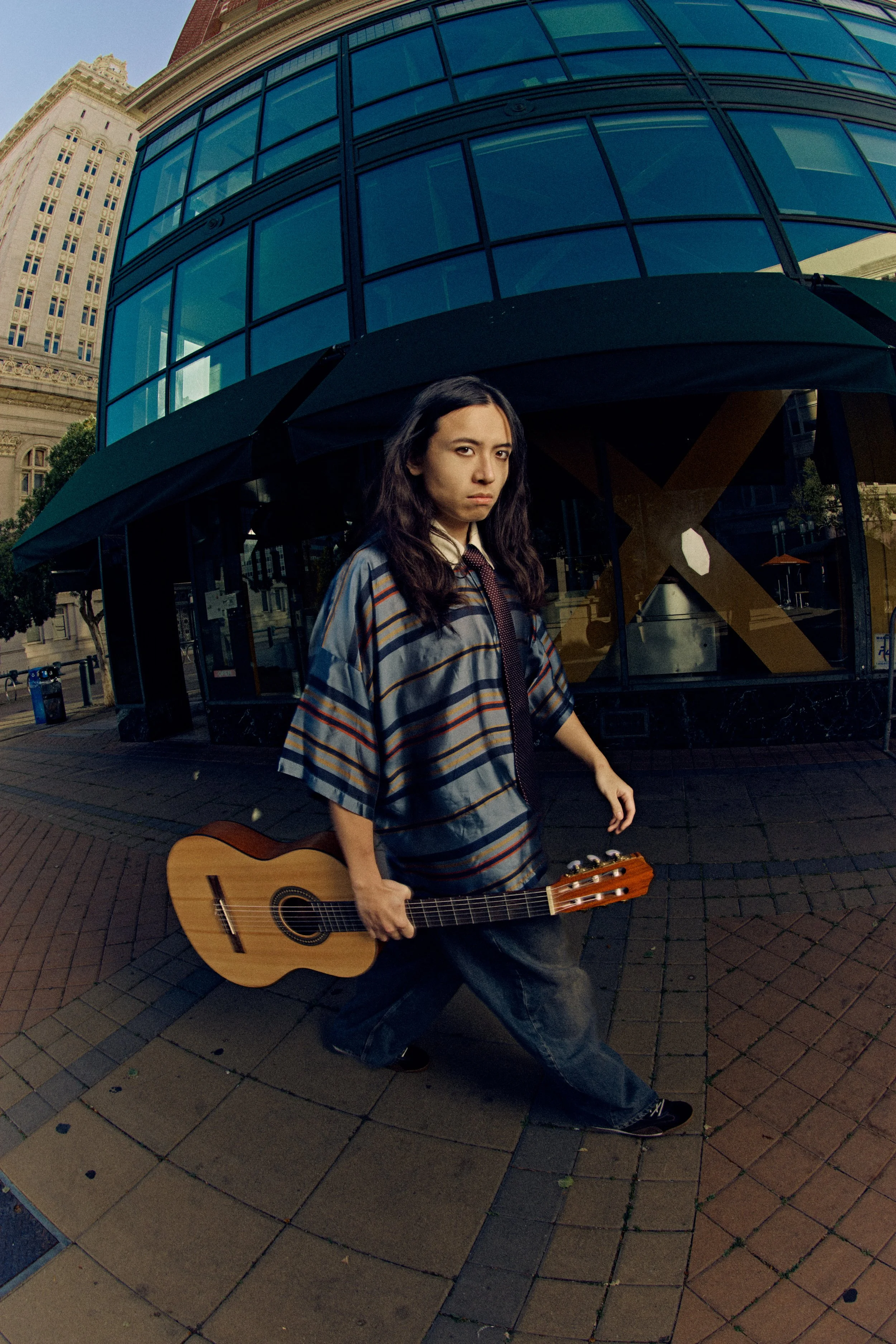 A person with long dark hair wearing a striped shirt and black pants holding a guitar, standing on a brick sidewalk in front of a modern glass building.
