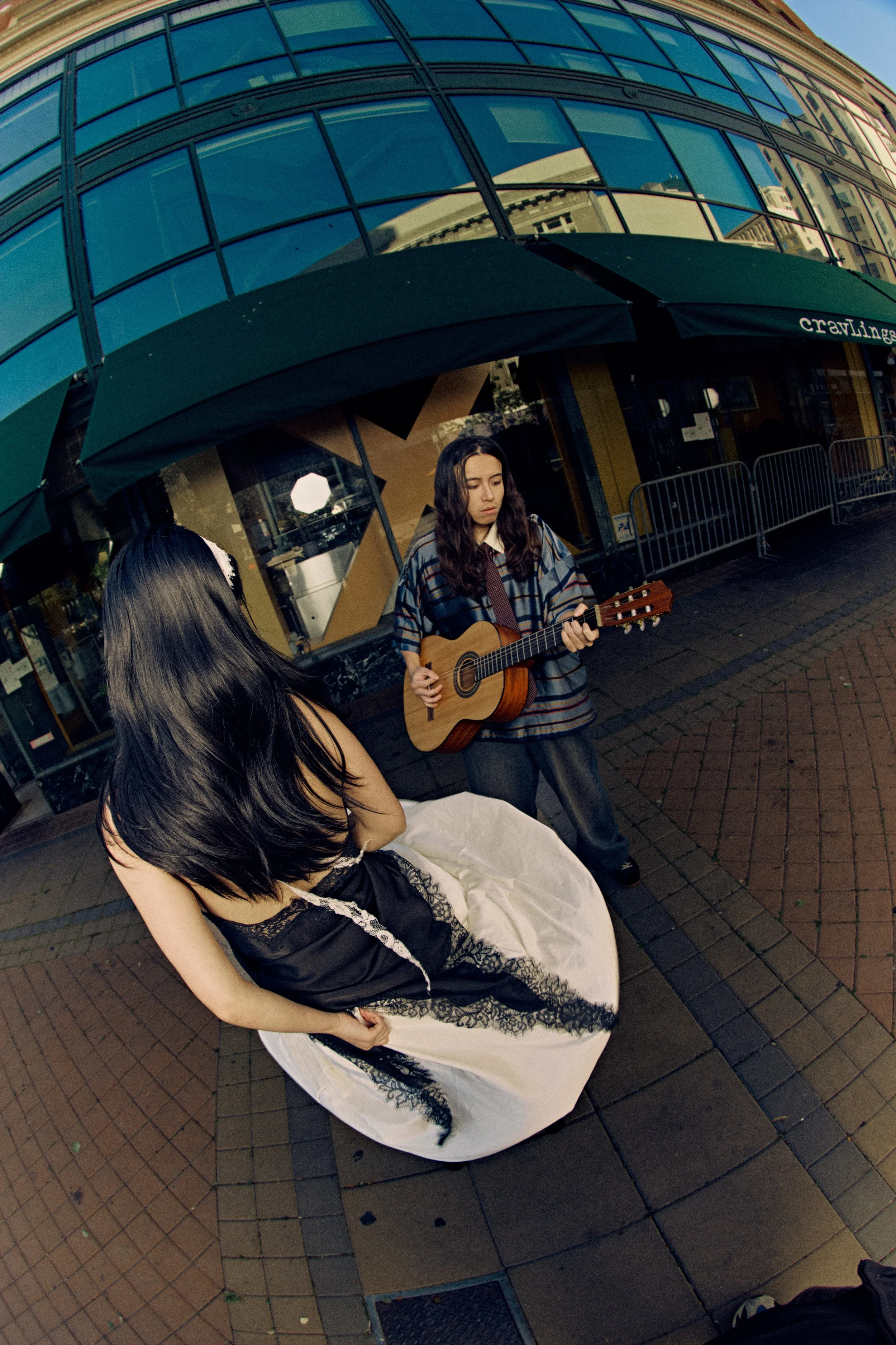 Two young women are outdoors, one standing in a wedding dress with black lace details and the other, with wavy brunette hair, playing a guitar. They are under a green umbrella in front of a modern building with glass windows.