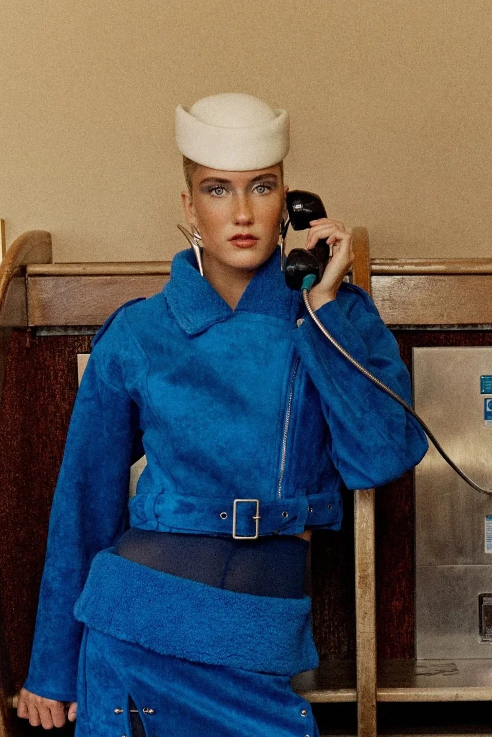 A woman with bright makeup wearing a white hat, large earrings, and a blue outfit, talking on a wall-mounted telephone.