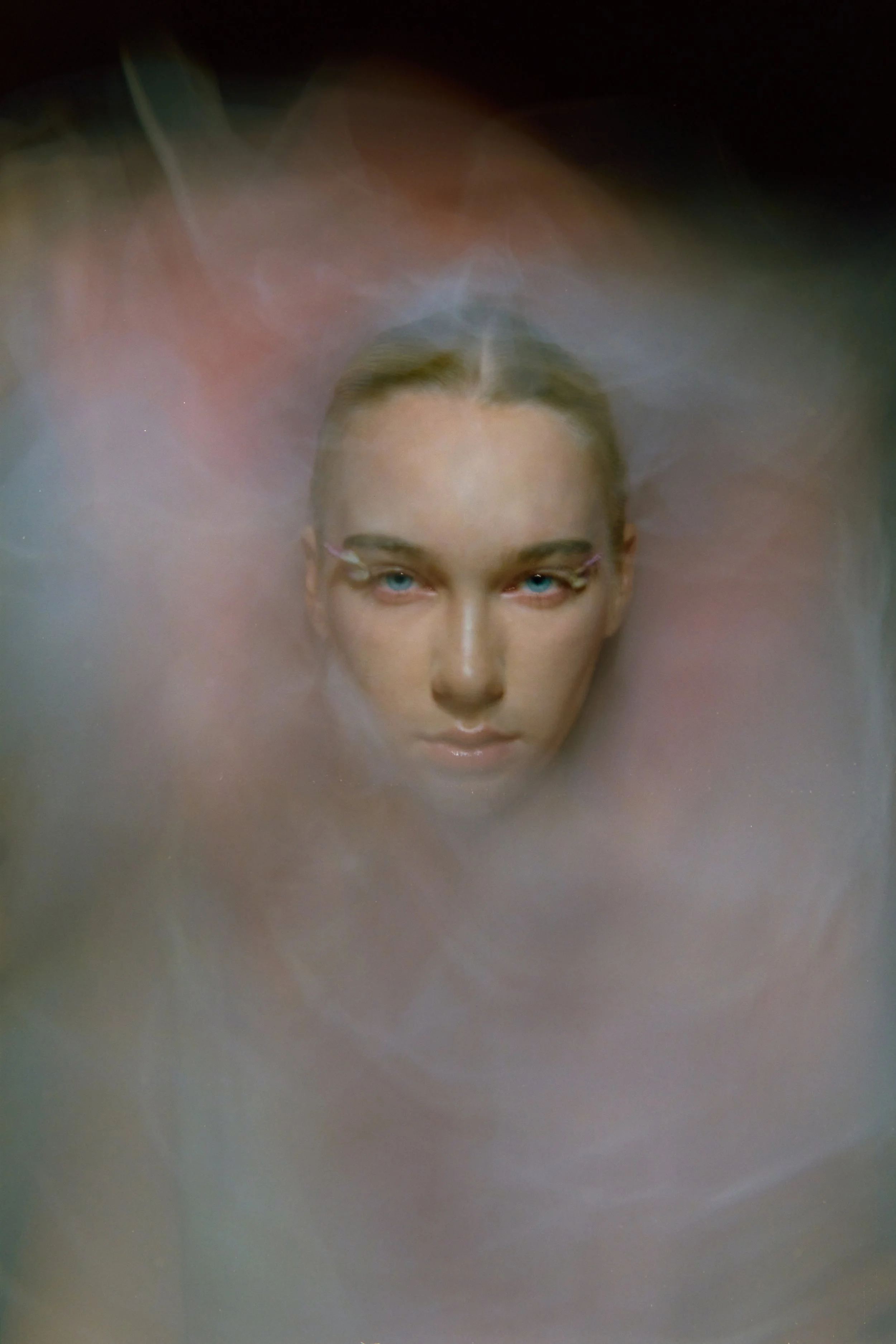 Close-up of a mannequin's head with blonde hair and blue eyes, partially submerged in a foggy, colorful environment.