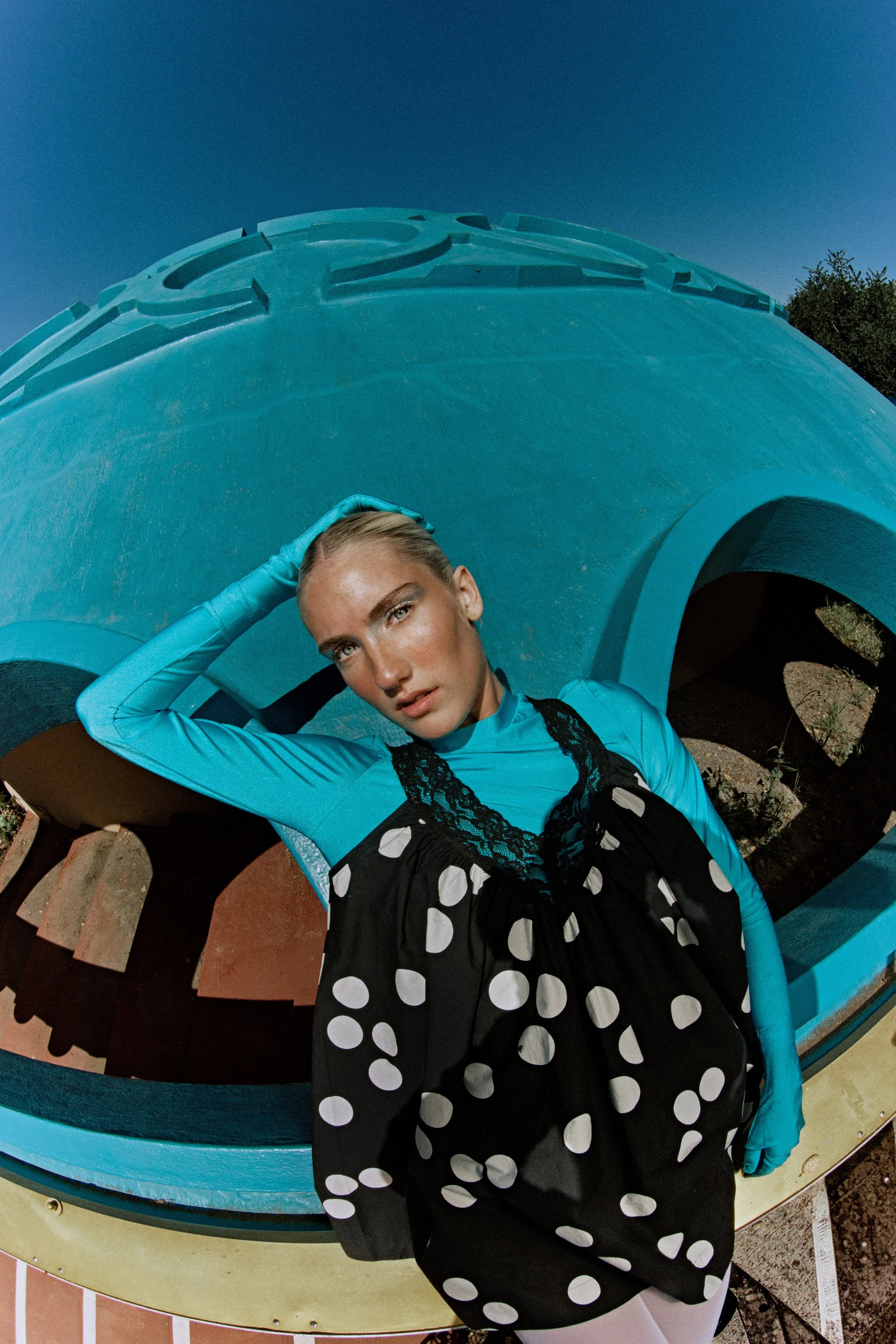 A woman with blonde hair, wearing a long-sleeved blue top with ruffled details and a black skirt with white polka dots, is posing in front of a large turquoise sculpture. She is touching her hair with one hand and looking at the camera. The sculpture