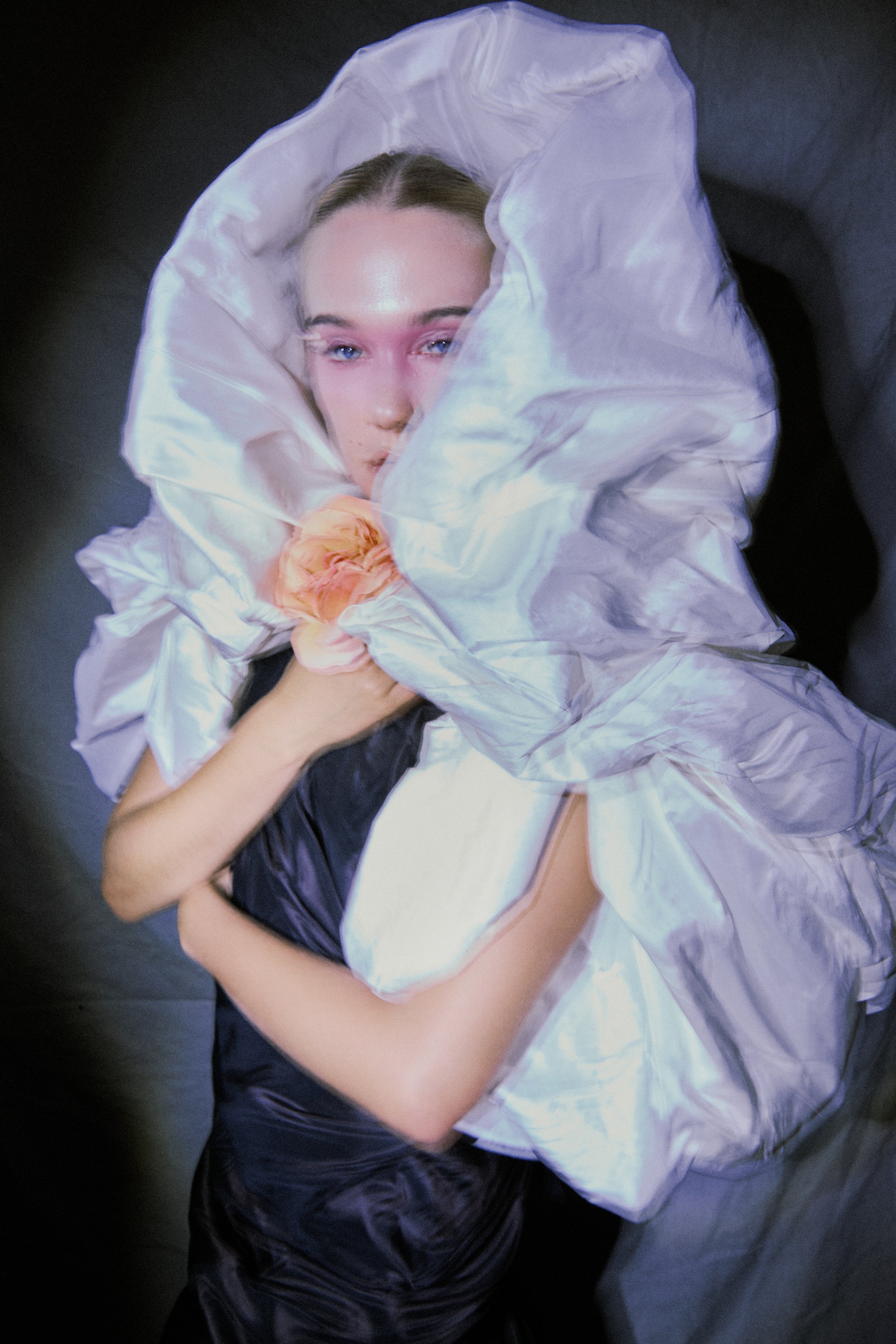 A woman lying down, partially wrapped in a large, shiny white fabric, with a peach-colored flower near her shoulder, staring directly at the camera with blue eyes.
