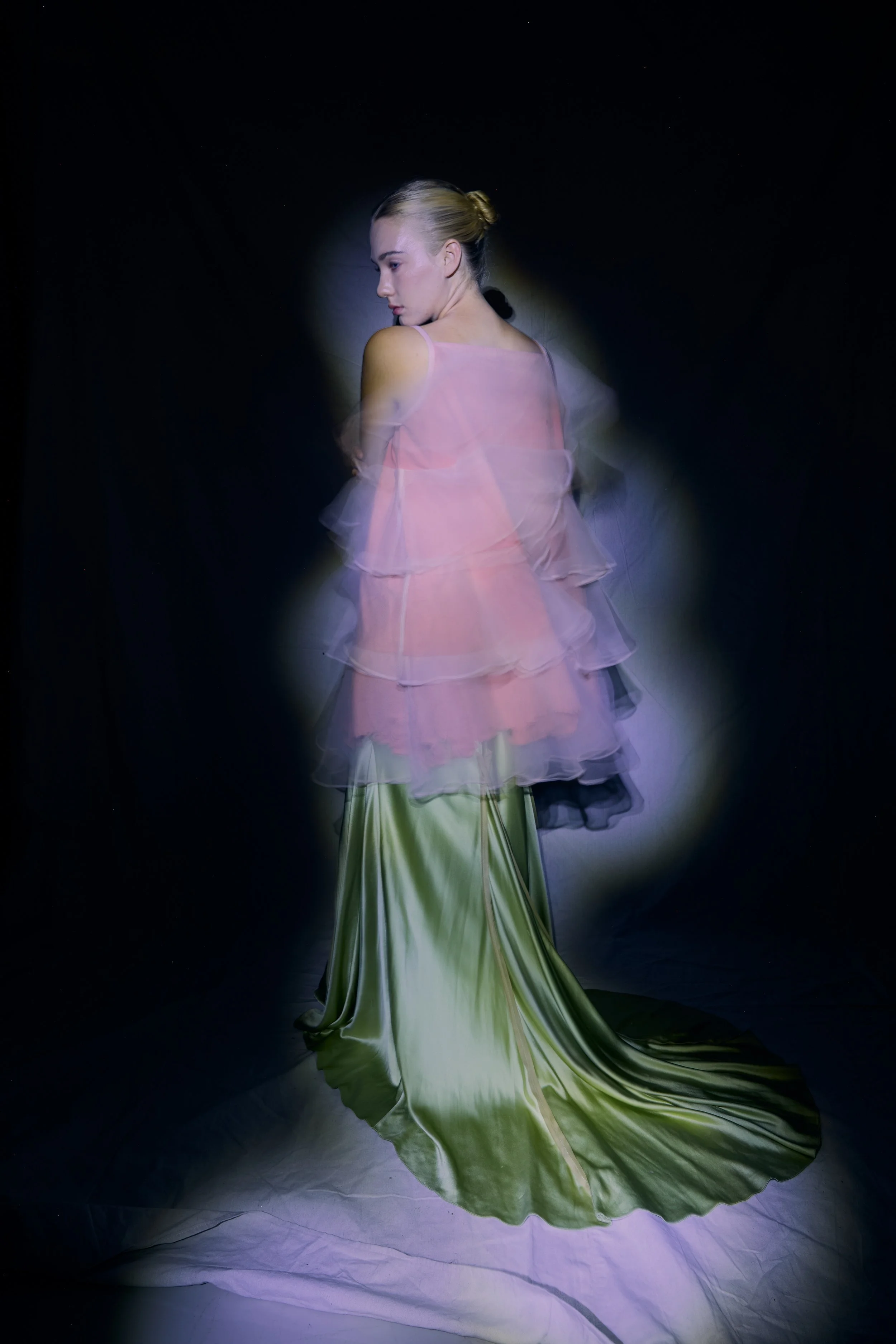 A young woman in an elegant, flowing green dress with a long train, standing against a dark background, illuminated by soft lighting.