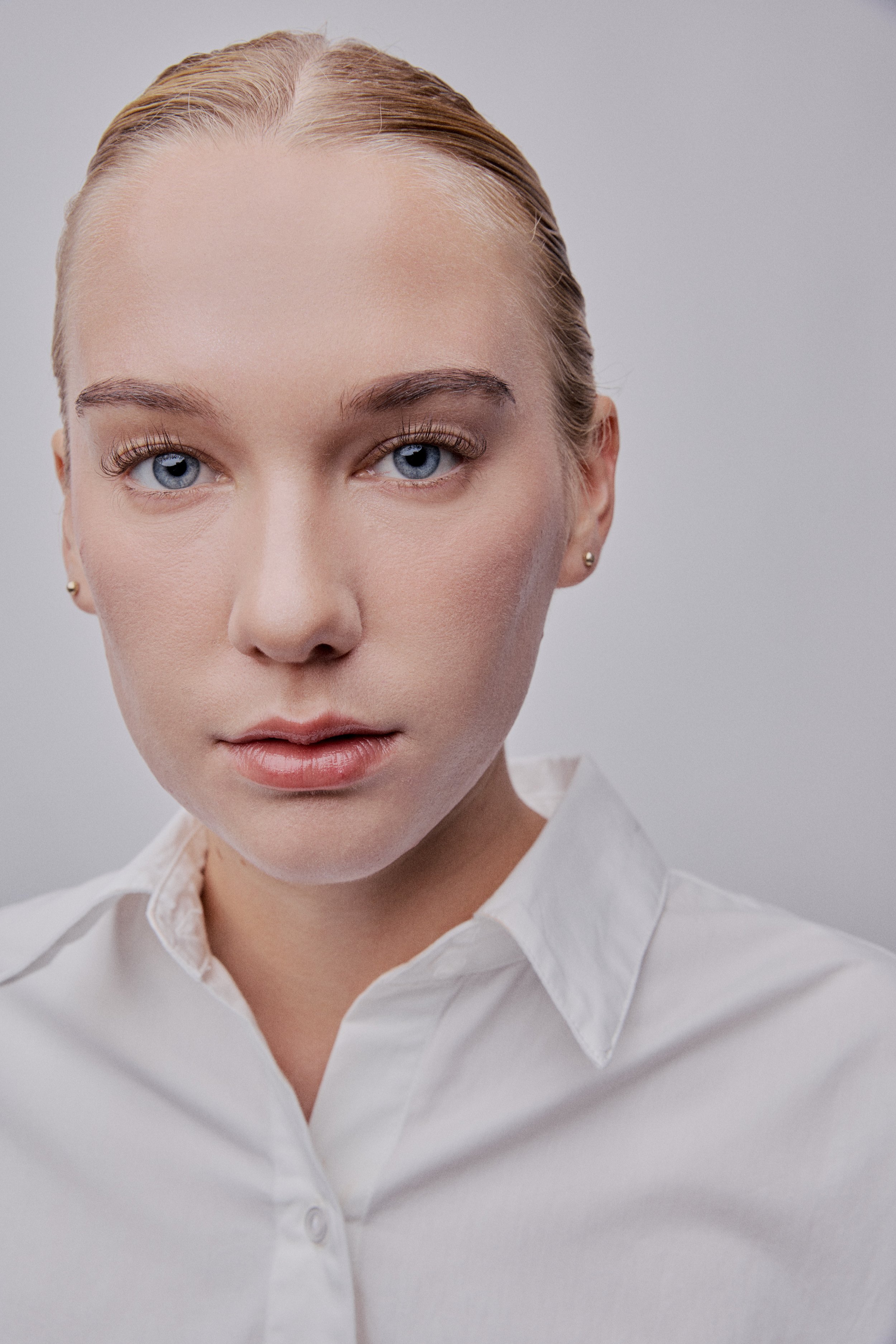 Close-up of a woman's face with blue eyes, blonde hair slicked back, wearing a white collared shirt, against a plain gray background.