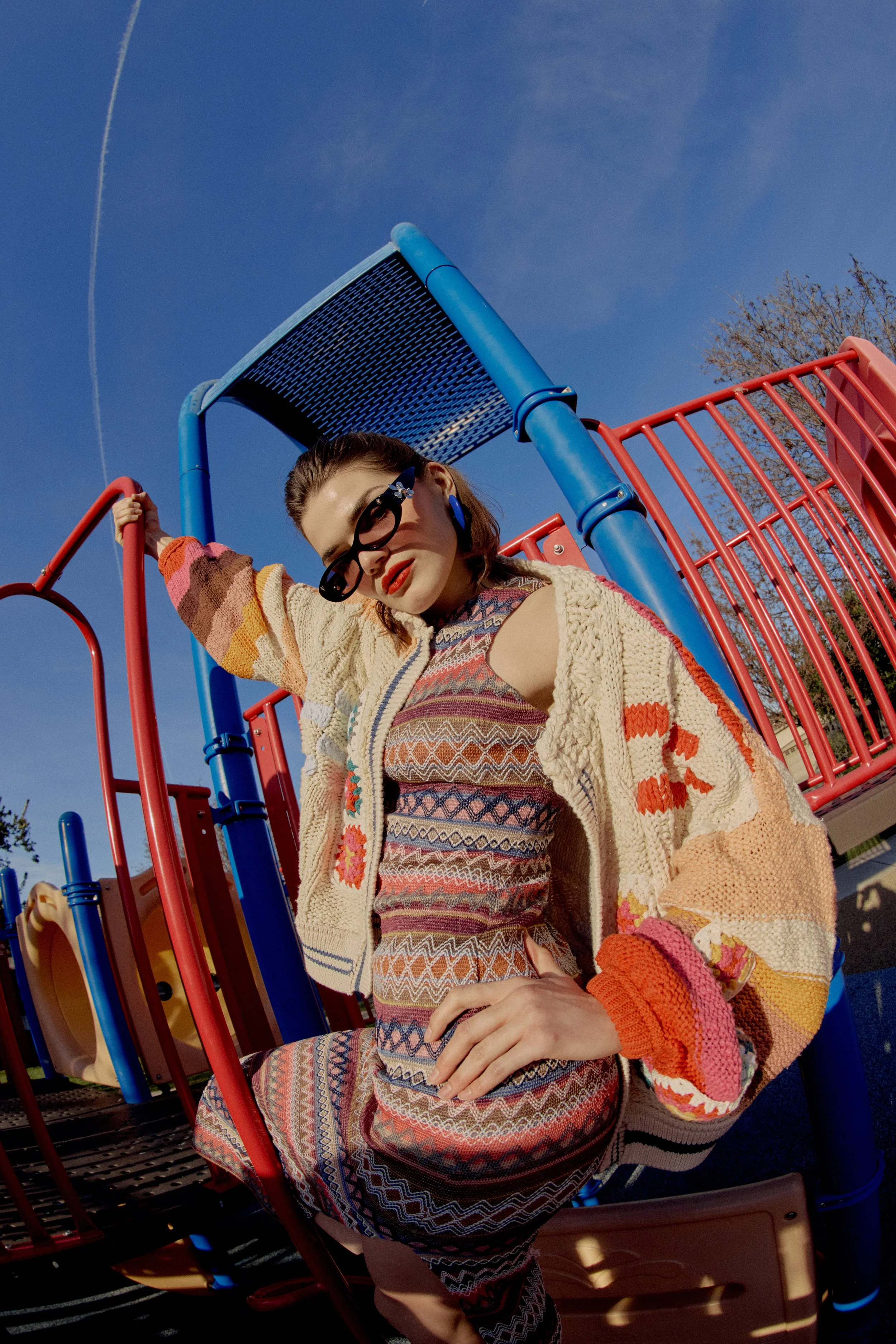 Young woman with dark hair, wearing sunglasses, a colorful patterned dress, and an oversized knitted cardigan, leaning on playground equipment with a blue sky in the background.