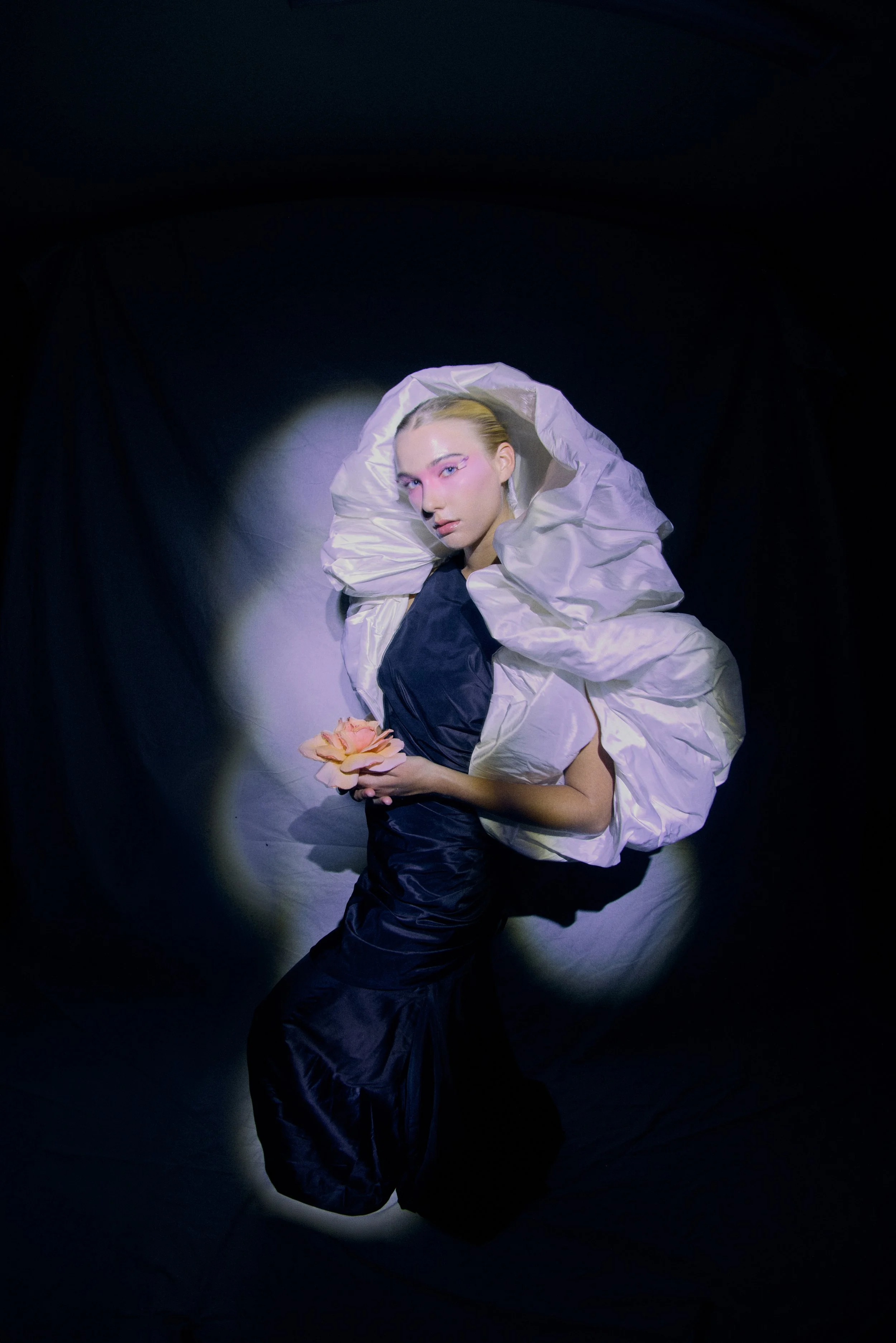 A woman with styled blonde hair and pink makeup poses in a dark setting, wearing a large, ruffled white collar and black satin dress, holding a pink flower.