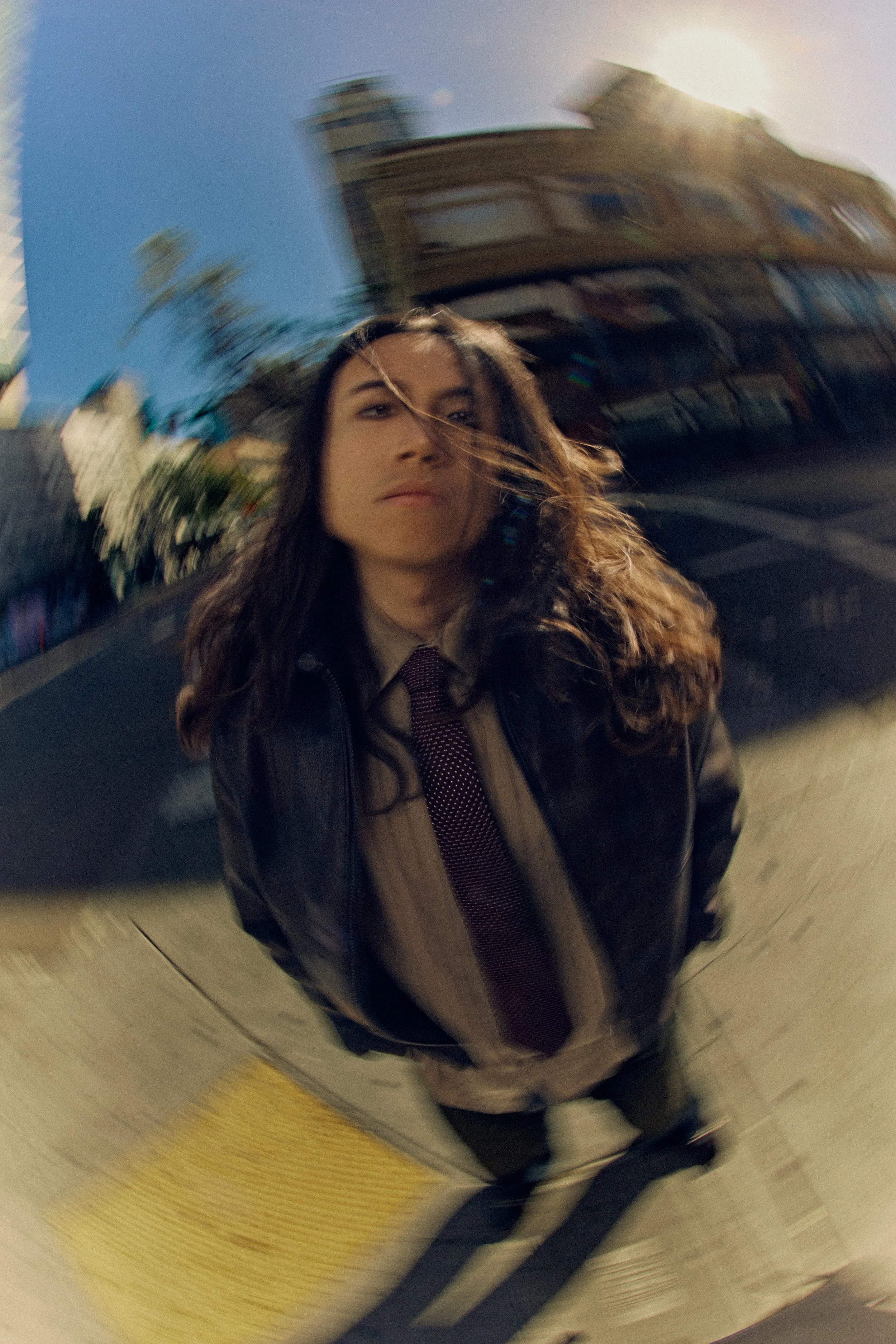 A person taking a photo with a fisheye lens, creating a distorted, circular effect. The person has long wavy hair, is wearing a leather jacket, and a tie, and is standing in front of buildings and trees during daytime.
