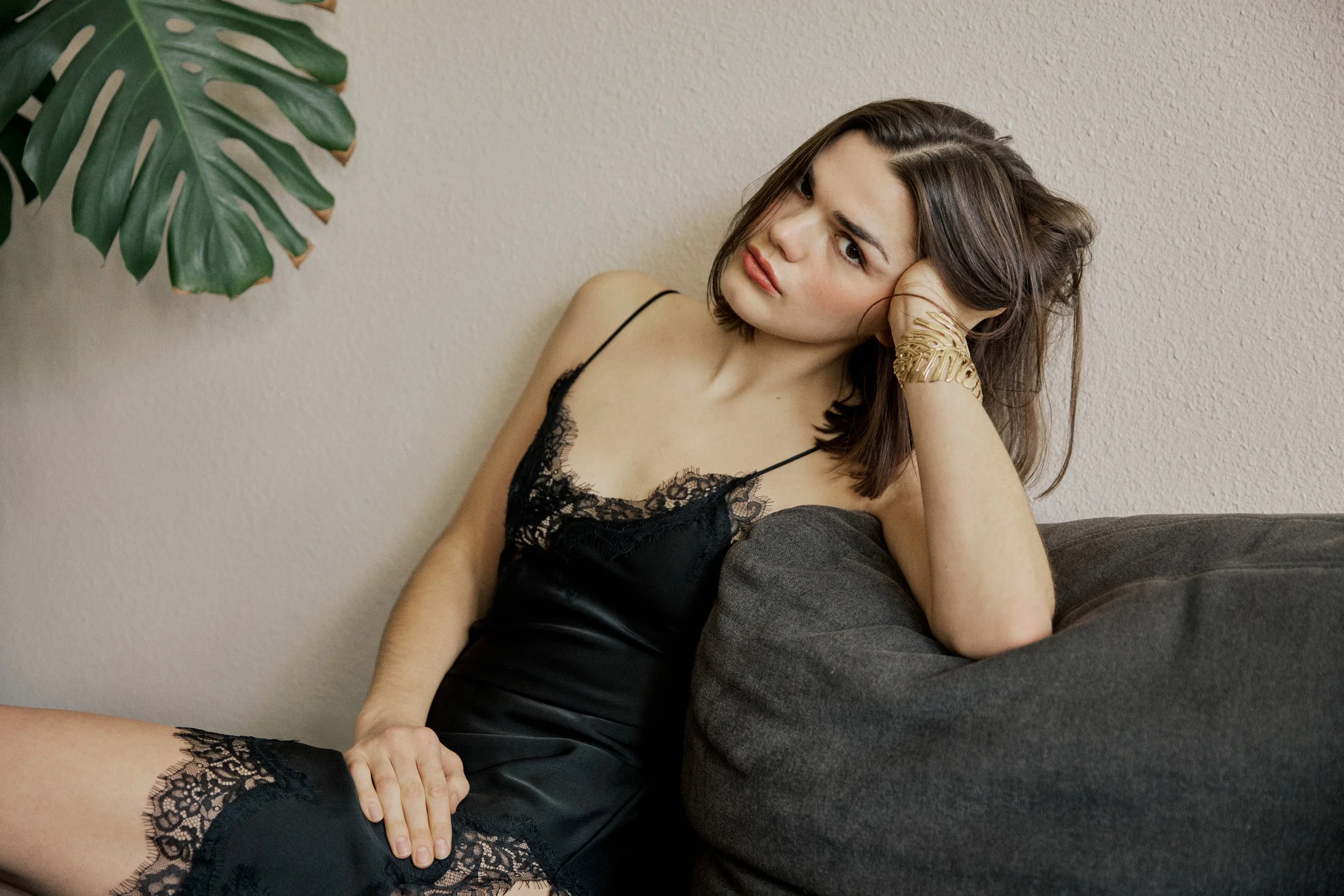 A young woman with shoulder-length brown hair sitting on a gray couch, wearing a black lace camisole, with one hand resting on her head and a large green monstera plant leaf visible in the upper left corner.