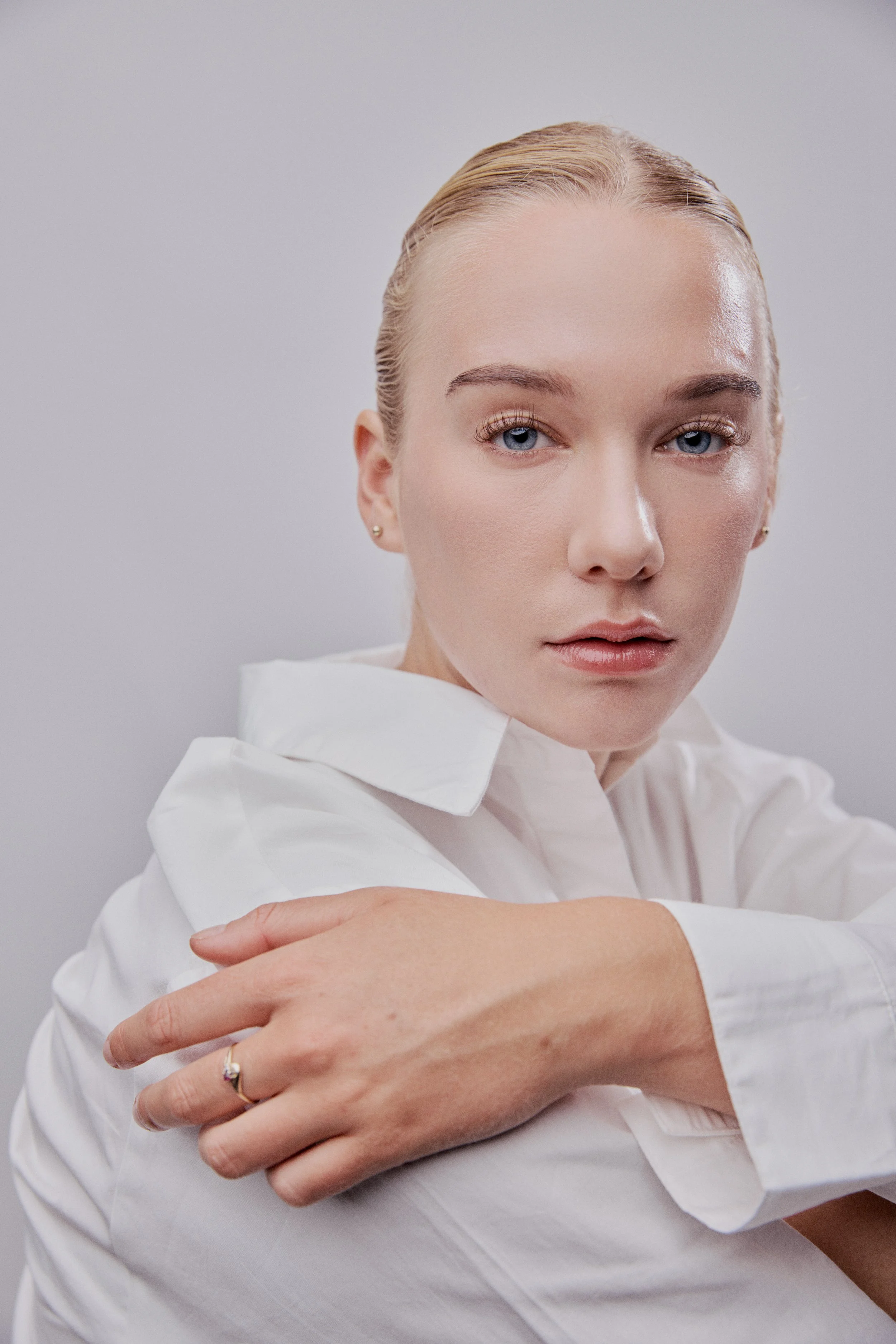 A woman with blonde hair and blue eyes wearing a white shirt, resting her arm on her knee, against a plain light background.