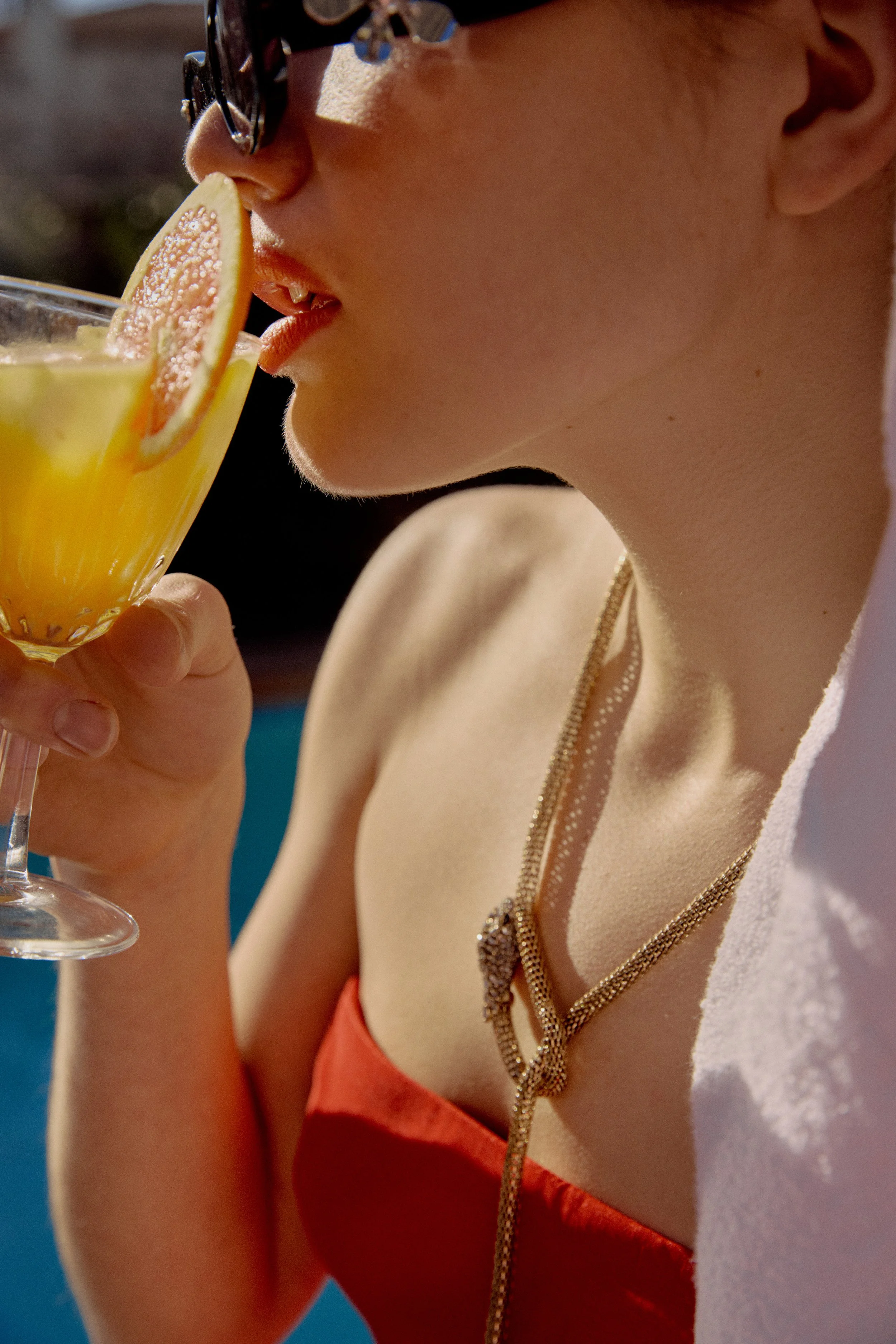 Close-up of a person wearing sunglasses with a beige dress with gold chains around the neckline, sipping a drink garnished with a slice of grapefruit.