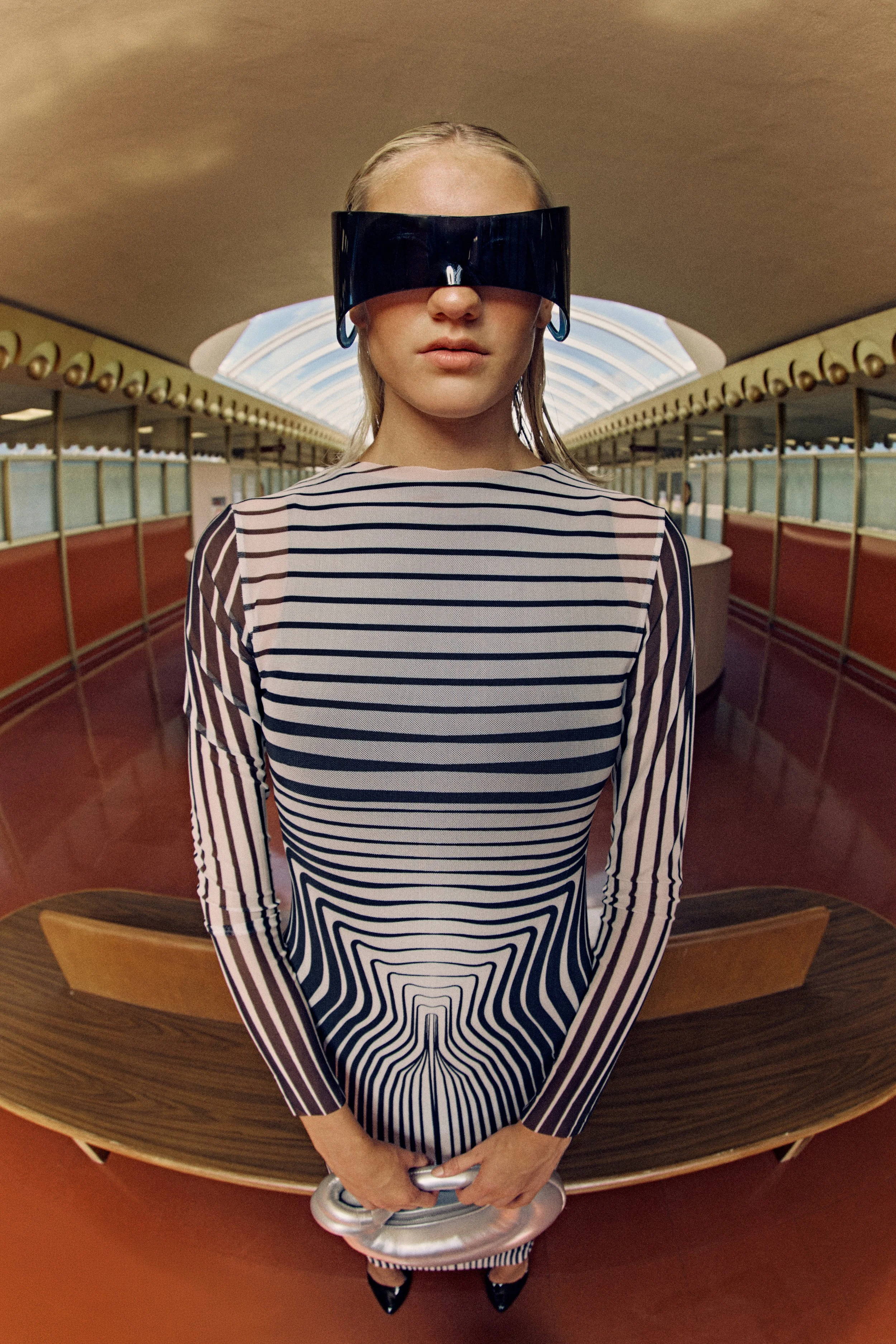 A woman with blonde hair wearing a striped dress and futuristic black visor glasses stands in a retro-style diner, holding a small robot or object, with a curved ceiling skylight and wood-paneled polka-dot wall in the background.