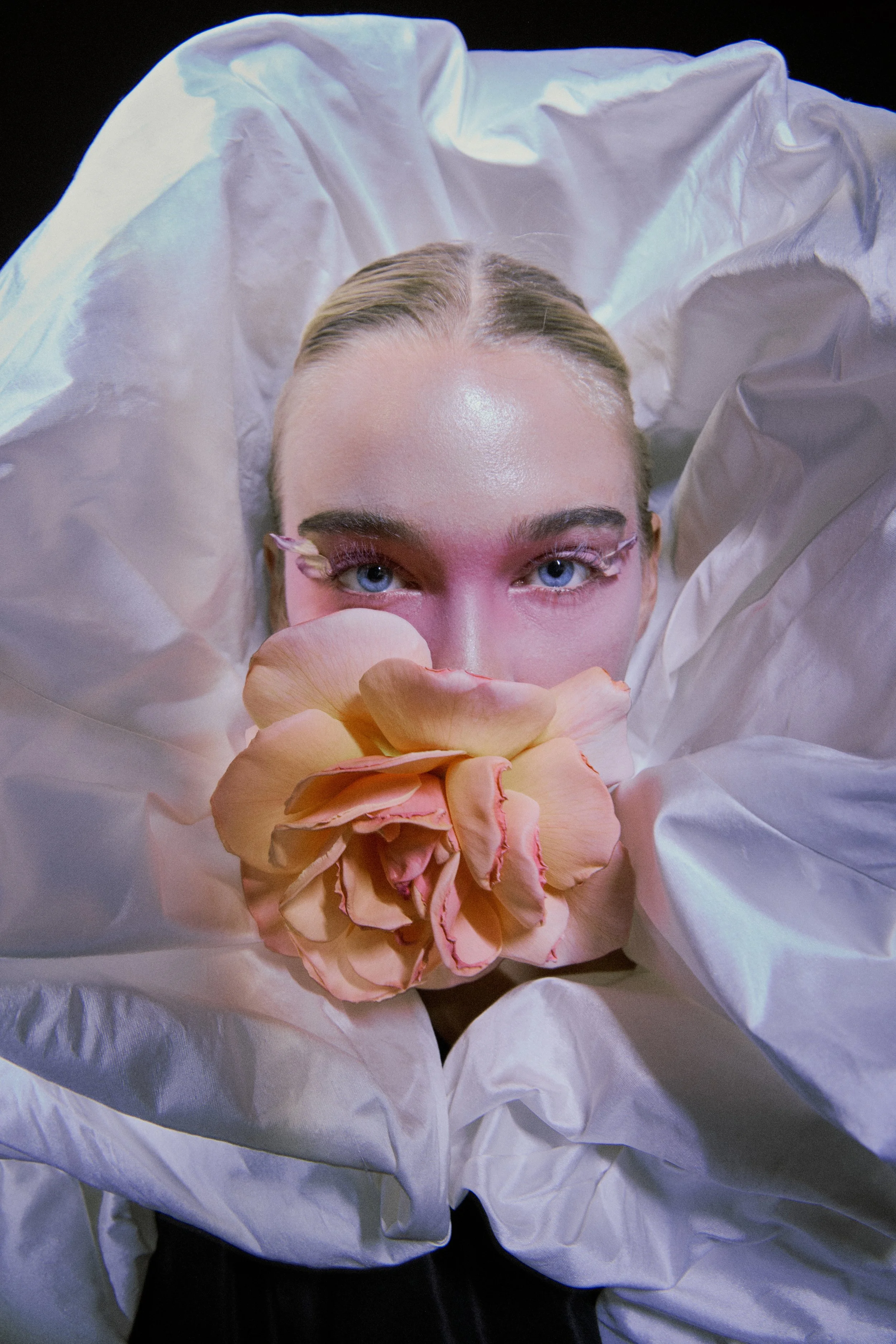 A woman with blonde hair and blue eyes, lying on a white sheet, holding a large pink and peach flower near her face, with a black background.
