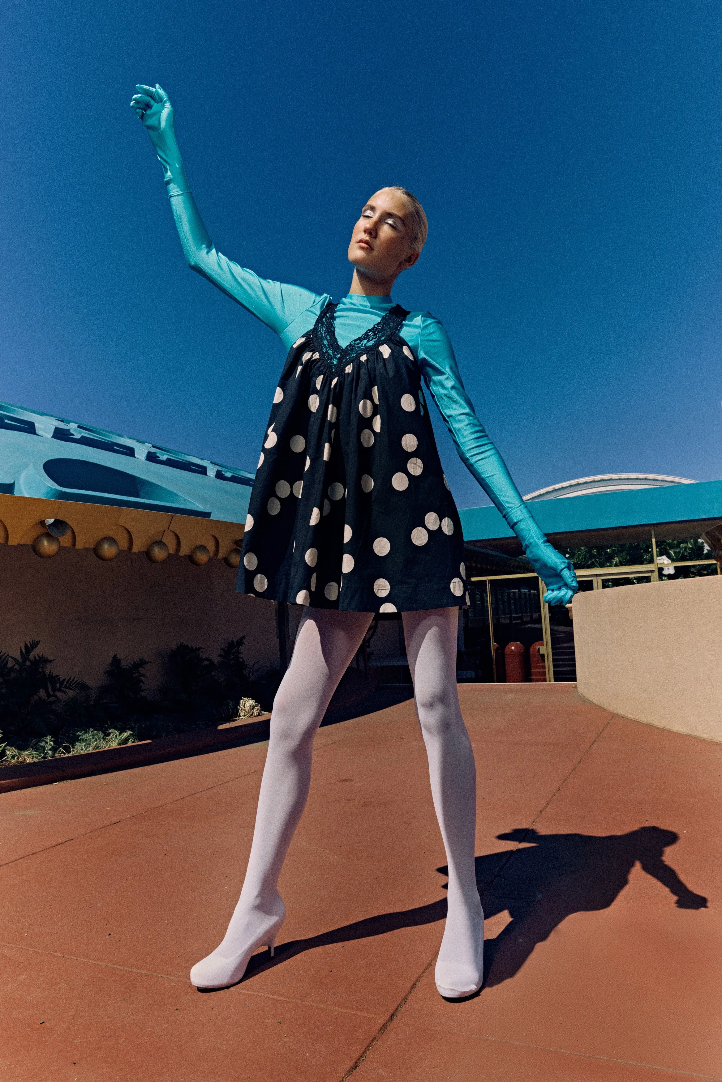 A person dressed in a retro style costume with white fishnet stockings, a polka dot dress, tight blue gloves, and light makeup poses with one arm raised and eyes closed against a clear blue sky. The setting appears to be an amusement park or a simila