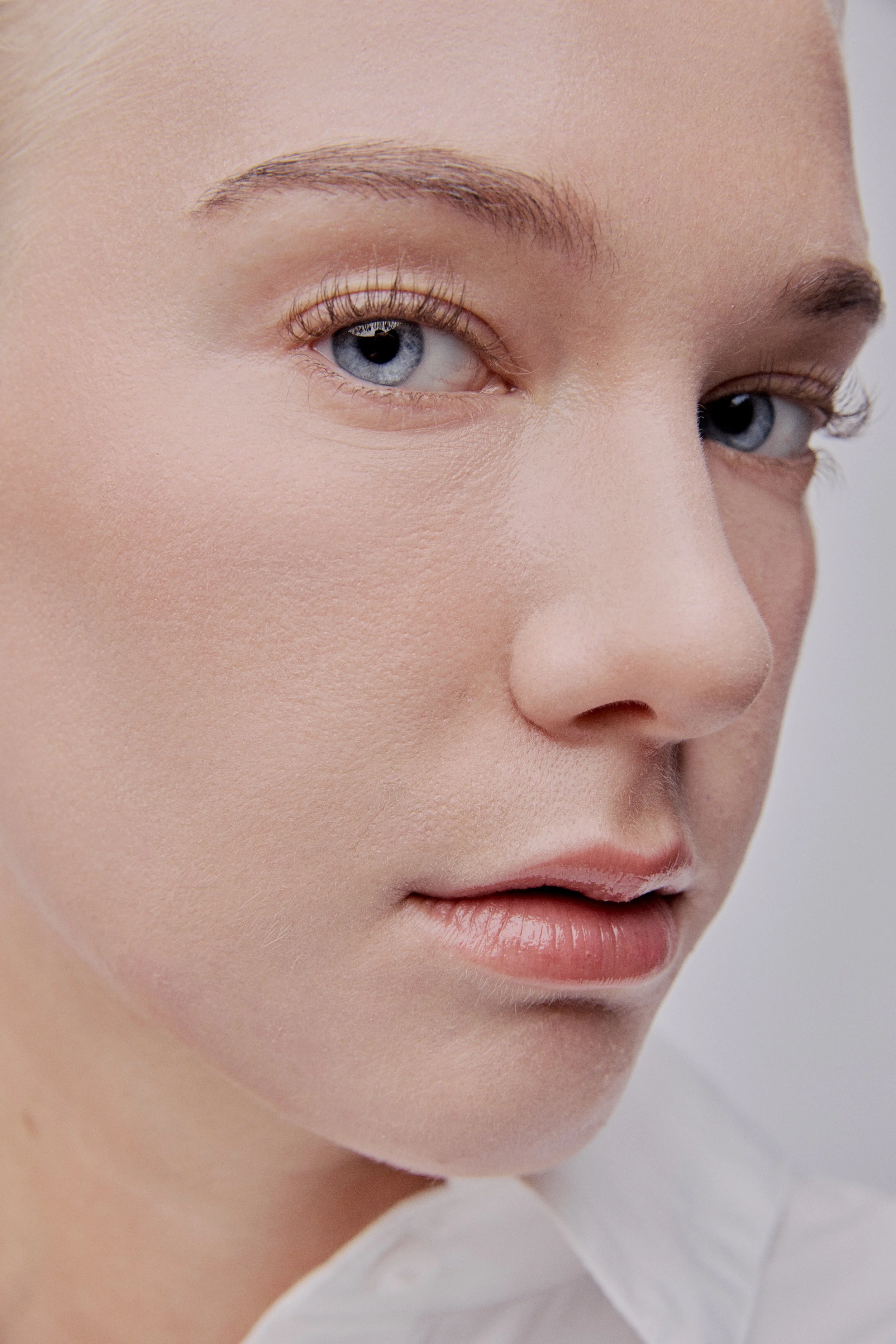 Close-up of a woman's face with fair skin, blue eyes, light makeup, and soft pink lips, wearing a white shirt.