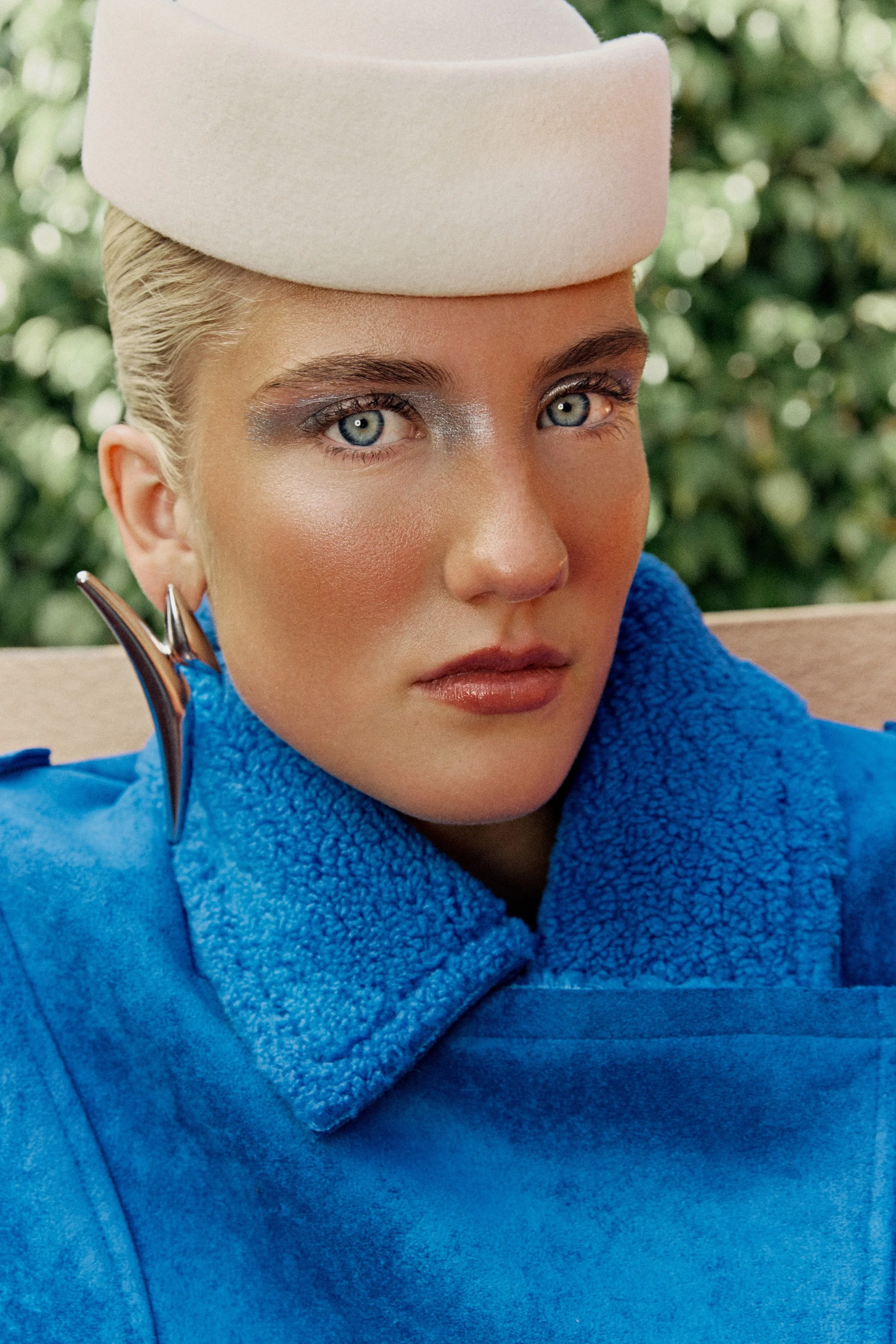 Close-up of a woman with platinum blonde hair, wearing a cream-colored hat, silver eye makeup, blue fleece jacket, and silver earrings, with greenery in the background.